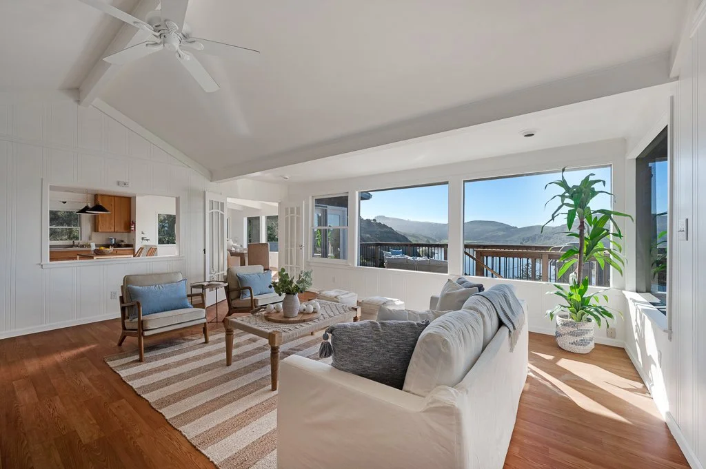 Living room with large windows overlooking hills, white walls, hardwood floor, white sofa, wooden coffee table with decor, two armchairs with blue cushions, large potted plant, ceiling fan, open kitchen in the background.