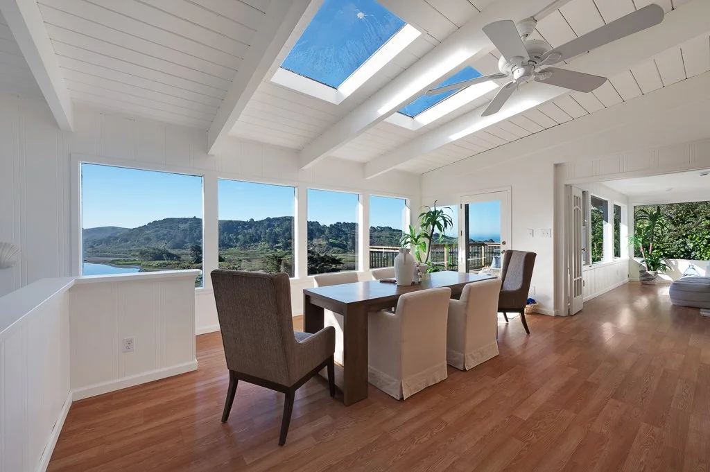 Bright dining room with large windows and skylights, wooden floor, white ceiling, ceiling fan, and plants, overlooking a scenic landscape with hills and water.