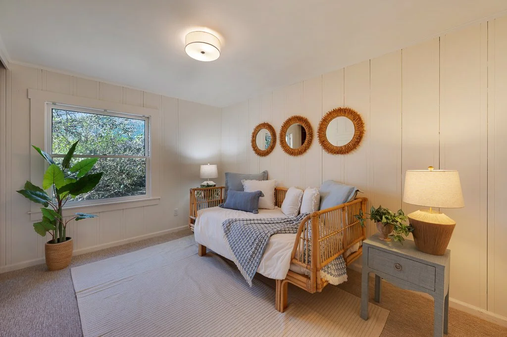 A cozy bedroom with white-paneled walls, a wooden daybed with white and blue bedding, a window with greenery outside, and a side table with a lamp and potted plants. There are three round mirrors with wicker frames on the wall above the bed.