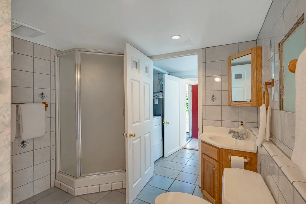 Bathroom with tiled walls and floors, a corner shower with a frosted glass door, a wooden framed mirror over the sink, a towel hanging on a wooden rack, a toilet with a toilet paper holder, and an open door leading to a laundry area and outside.