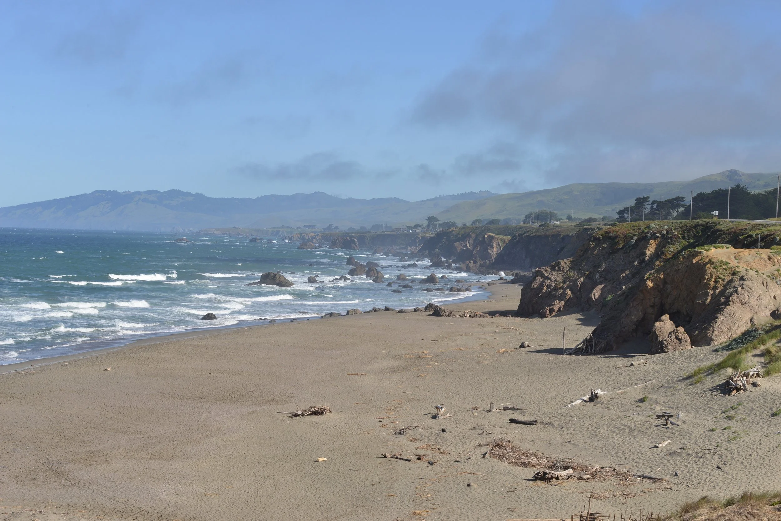 Sunny beach with sandy shore, rocks, and cliffs overlooking the ocean, with distant hills and scattered clouds in the sky.