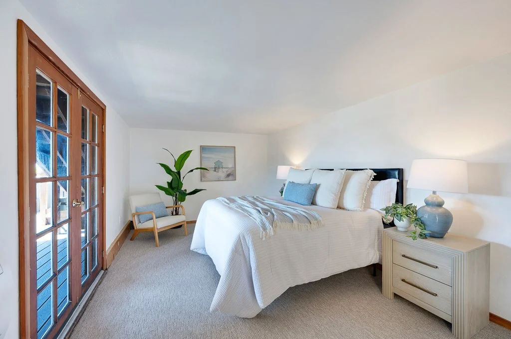 A cozy bedroom with a large bed, white bedding, and decorative pillows, next to a wooden nightstand with a lamp and vase. There is a sliding glass door with wooden frames, a small armchair with a cushion, and a large potted plant in the corner. A framed picture hangs on the wall.