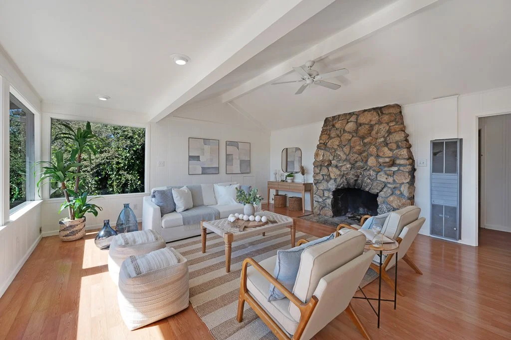 Living room with white walls, a stone fireplace, beige armchairs, a sofa, a wooden coffee table, various vases, and a large window with greenery outside.