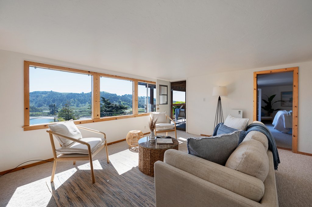 Living room with large windows offering scenic mountain views, furnished with a beige couch, white armchairs, a round wicker coffee table, a floor lamp, and a patterned rug, leading to a bedroom.