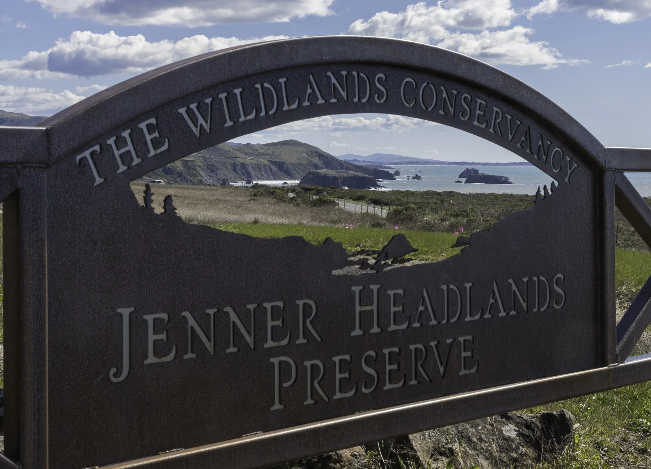 Sign for the Jenner Headlands Preserve with a background view of a coastline, cliffs, and the ocean under a partly cloudy sky.