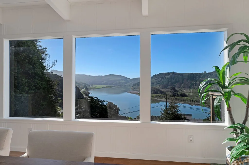 View of a lake with surrounding hills seen through large windows in a bright room, with a green potted plant on the right.
