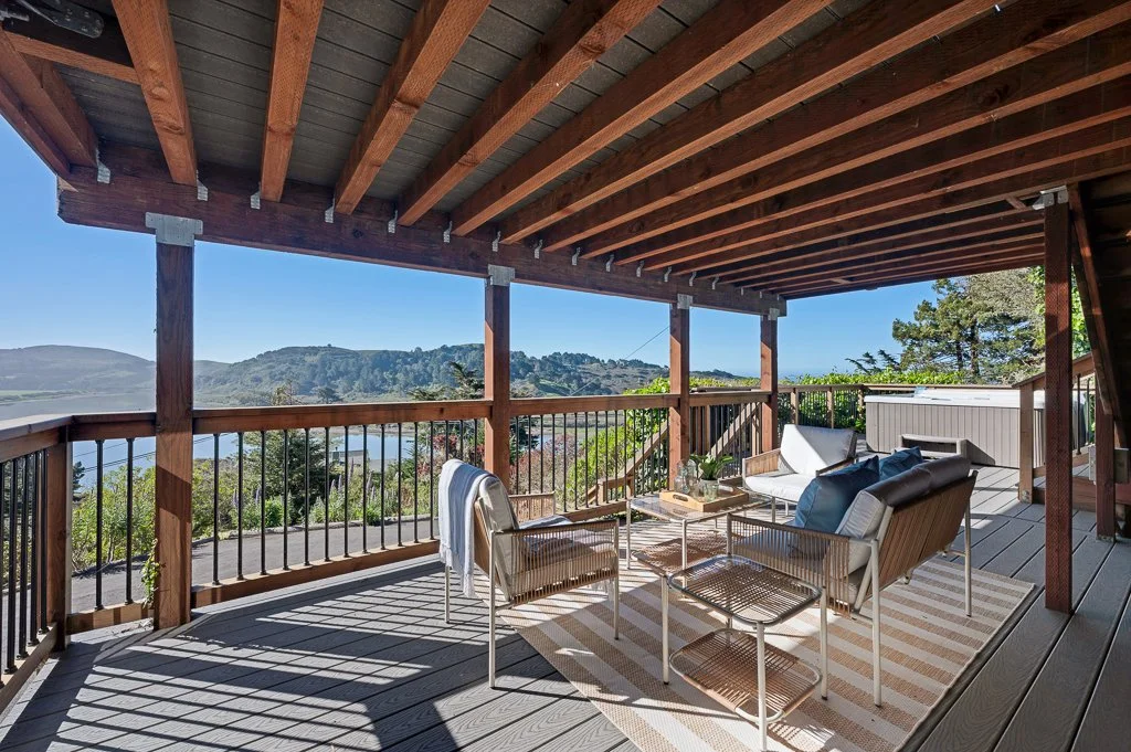 Outdoor balcony with wooden ceiling and railing, overlooking a hilly landscape with trees and water, furnished with chairs, cushions, a coffee table, and decorative items.