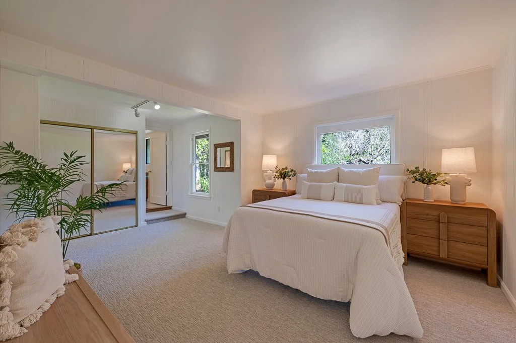 A bedroom with a large bed covered in white bedding, flanked by two wooden nightstands with lamps and plants, a window behind the bed, and a mirrored closet door in an airy, minimalist design.