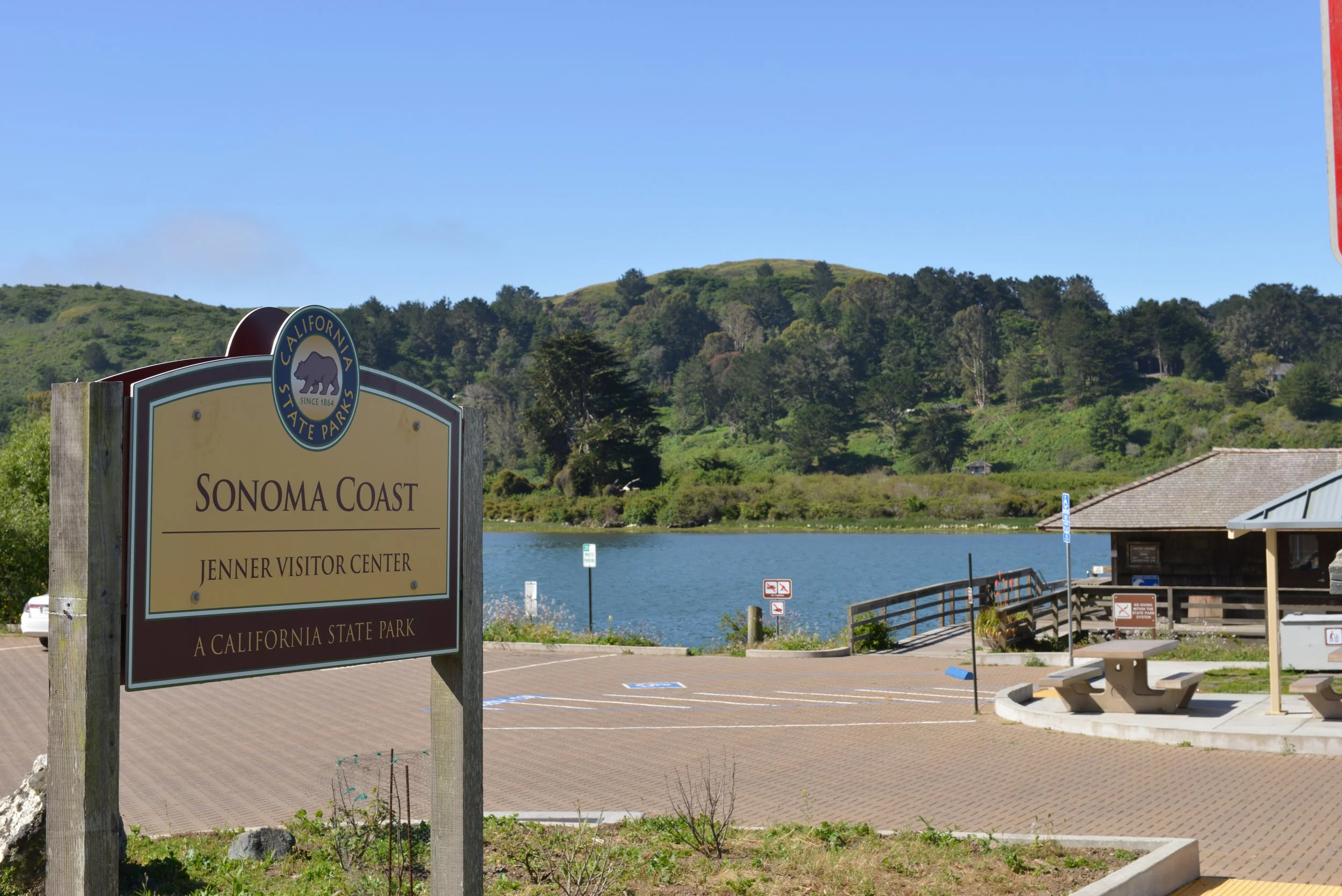 Sign at Sonoma Coast Jenner Visitor Center, California State Park, near a body of water with green hills and trees in the background and a clear blue sky.