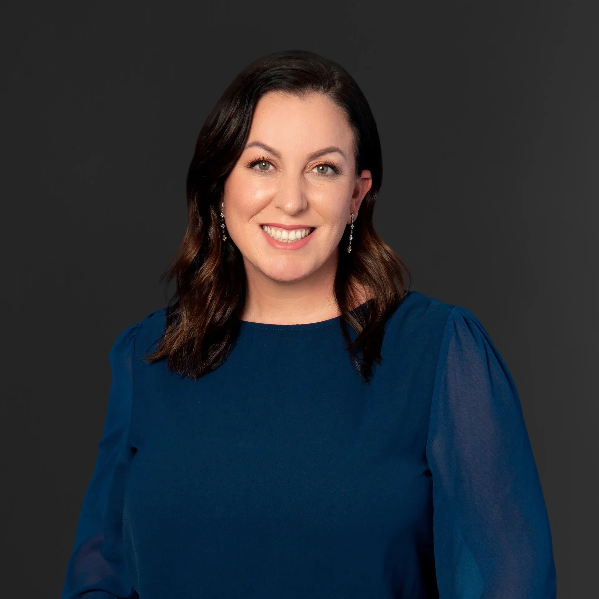 A woman with shoulder-length dark brown hair, blue eyes, and a bright smile, wearing a blue dress with puffed sleeves and dangling earrings, posed against a dark gray background.