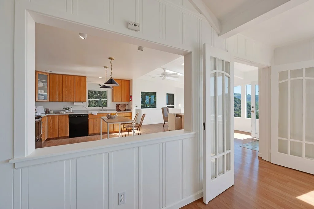 View of a bright, airy living space with a kitchen featuring wooden cabinets, a small dining table, and a living area with windows showing a green outdoor landscape.