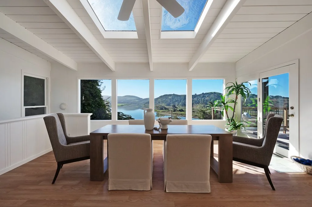 Bright dining room with large windows and skylight, wooden table with chairs, potted plant, and view of hills and water outside.