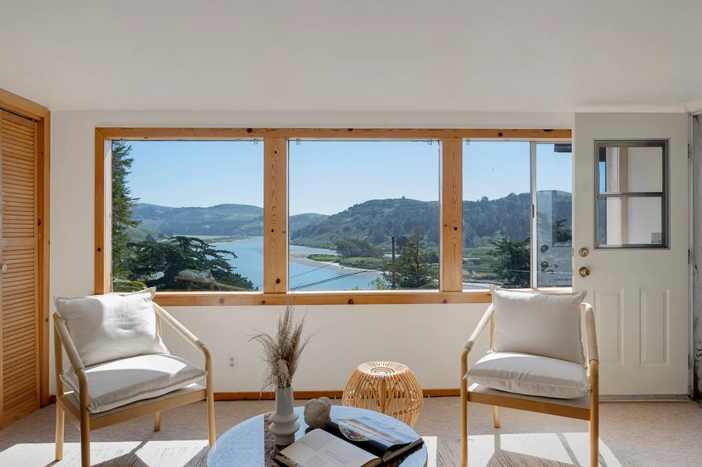 Living room with large window overlooking a river and hills, two white armchairs, a round coffee table with a book and a decorative vase, and a small woven stool.