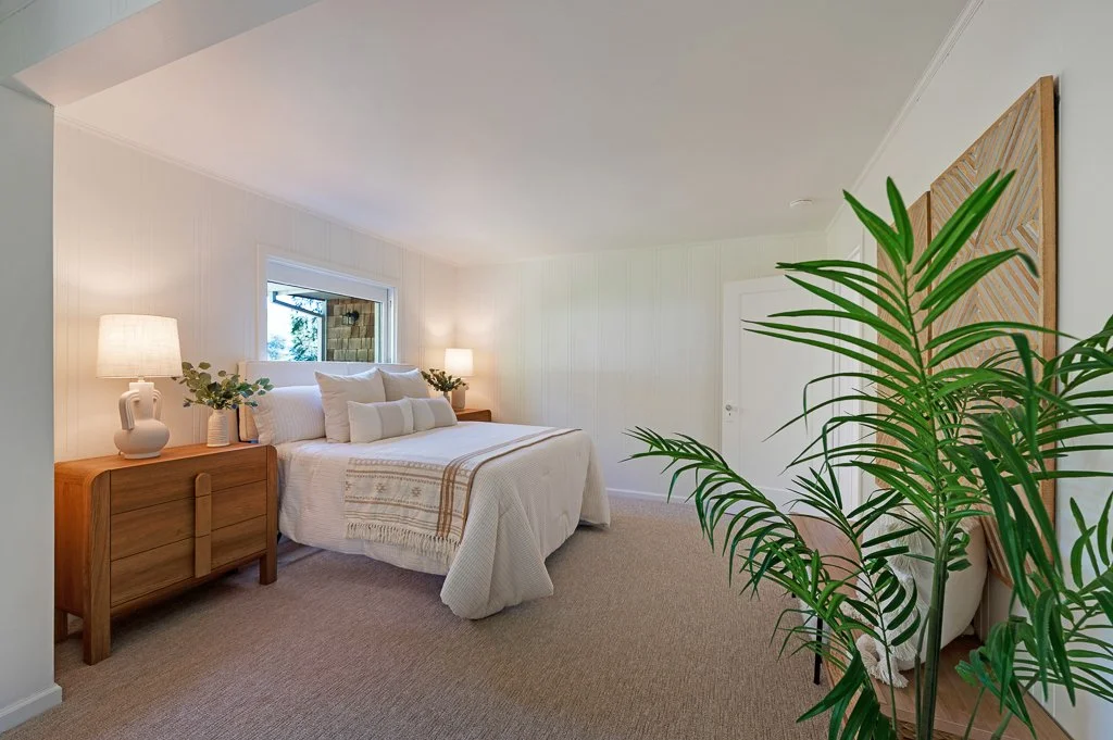 A cozy, well-lit bedroom with a large bed, side tables with lamps, decorative pillows, a window behind the bed, and a large green plant in the foreground.