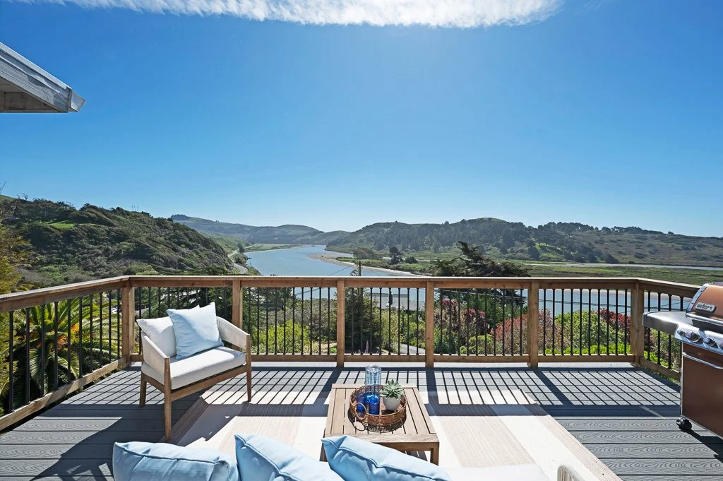 A wooden balcony overlooking a river and rolling hills, with seating and a barbecue grill.