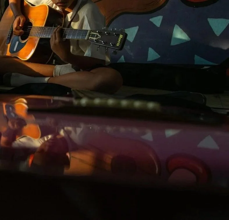 Ein Junge spielt auf einer Gitarre, während er auf einem Boden sitzt, im Hintergrund sind Tapeten mit geometrischen Mustern sichtbar.