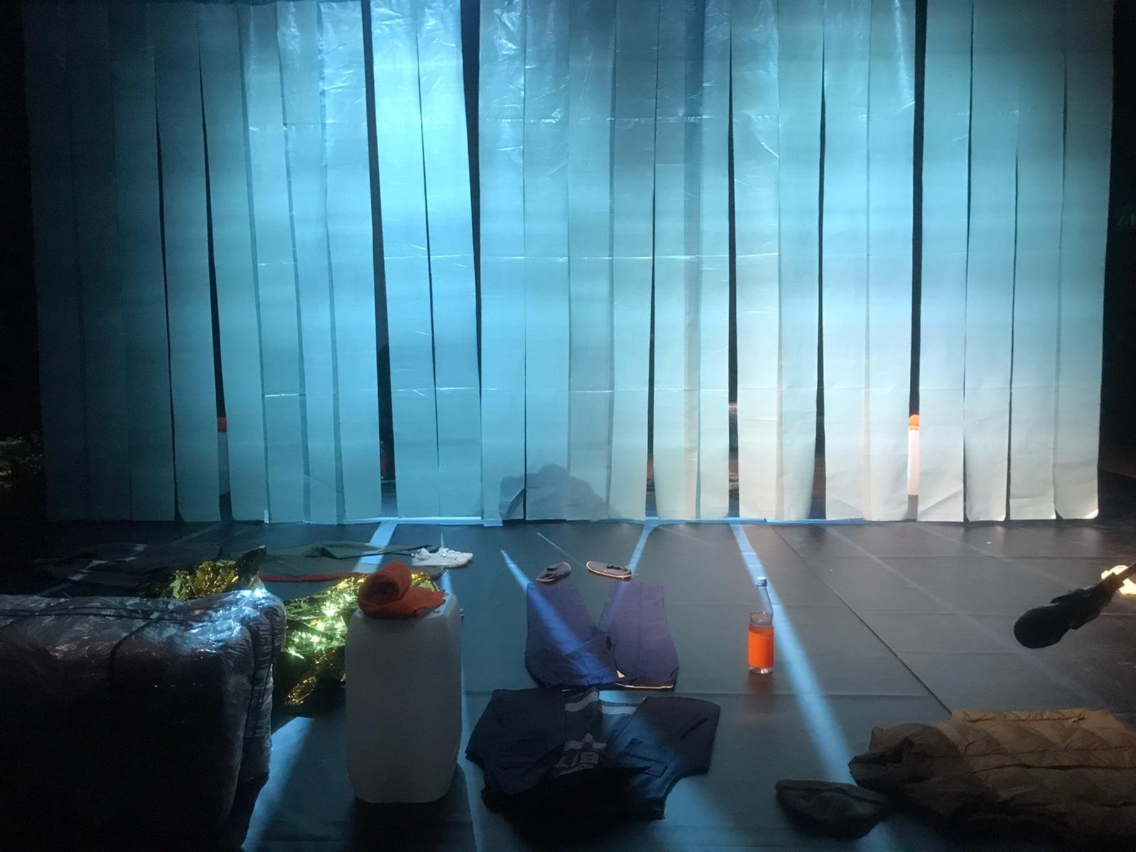 Eine dunkle Szene mit blauen Vorhängen und verstreuten Gegenständen auf dem Boden, darunter Schuhe, Wasserflaschen, Kleidung, Taschen und eine aufgerollte Matte.