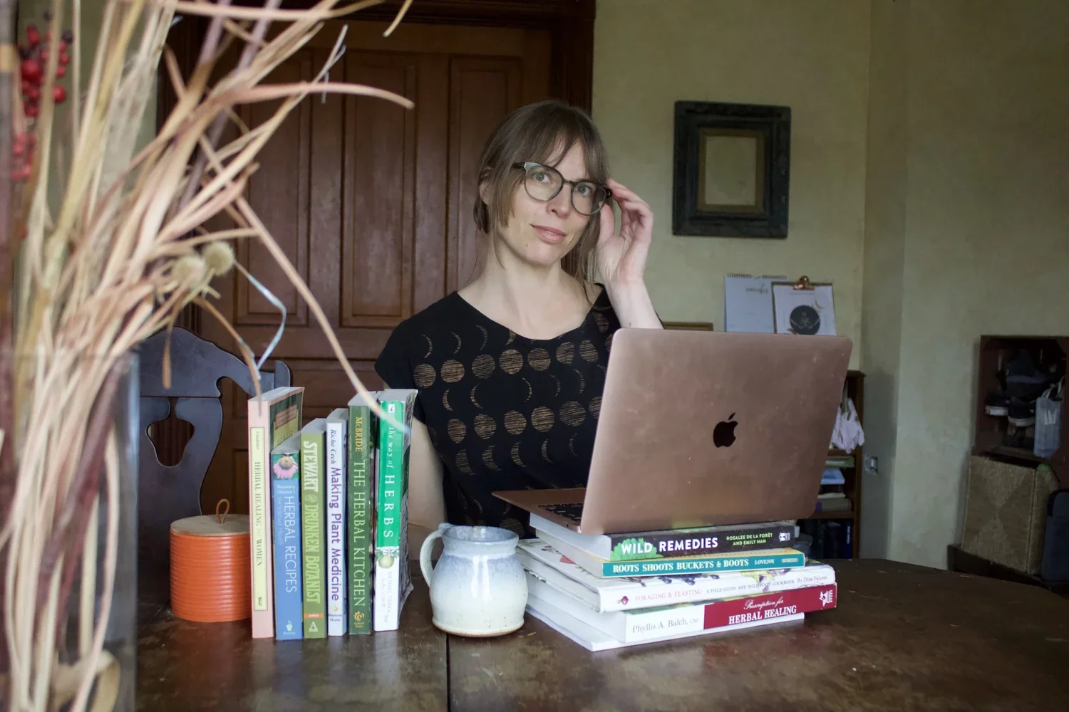 A woman with glasses sitting at a wooden dining table with a pink Apple MacBook in front of her. The table has a stack of books, a ceramic pitcher, and an orange container. She is adjusting her glasses, looking at the camera. The background features a wooden cabinet, a yellow wall, a framed picture, and miscellaneous items.