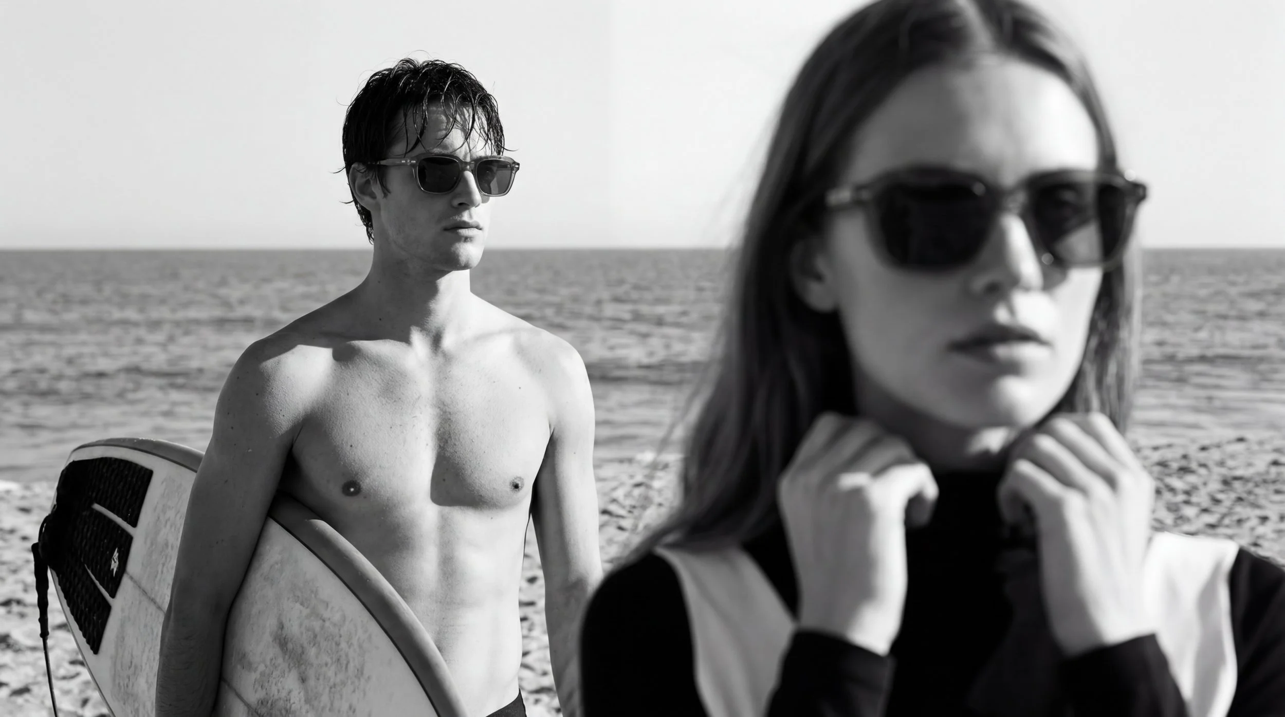A black and white photo of a shirtless man with sunglasses holding a surfboard, and a woman in sunglasses and a wetsuit near the ocean.