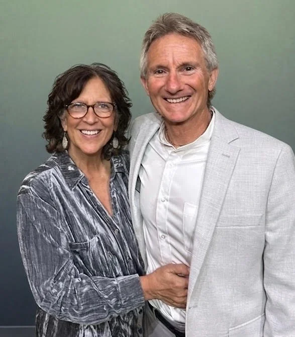 A smiling woman with curly dark hair and glasses, dressed in a striped shirt, standing next to a smiling man with gray hair, dressed in a light gray blazer, against a green background.