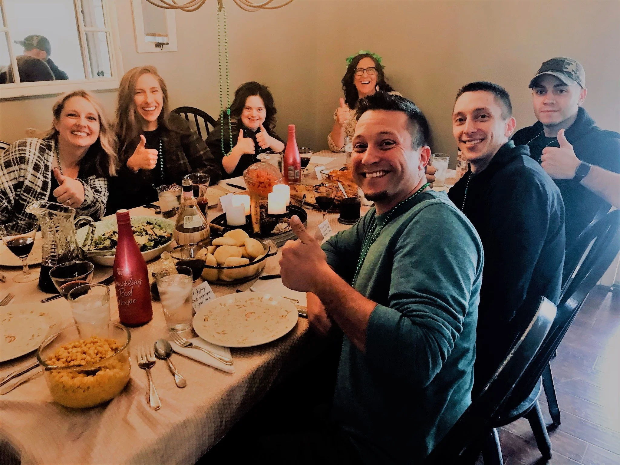 Group of eight people around a dining table, smiling and giving thumbs up, with food and drinks, during a celebration with candles and decorations.