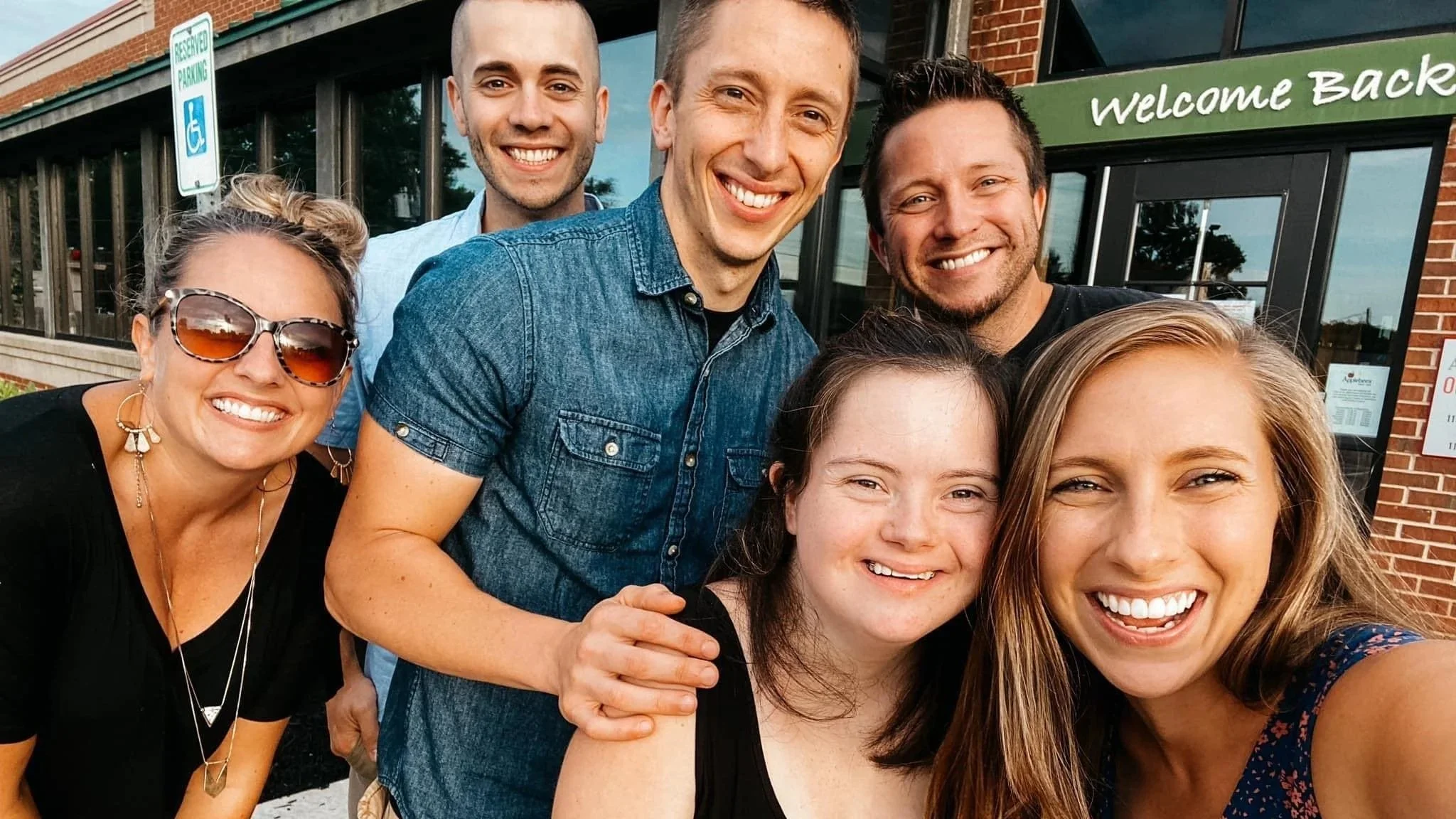 Group of seven smiling people taking a selfie in front of a building with a green sign that says "Welcome Back" and a reserved parking sign.