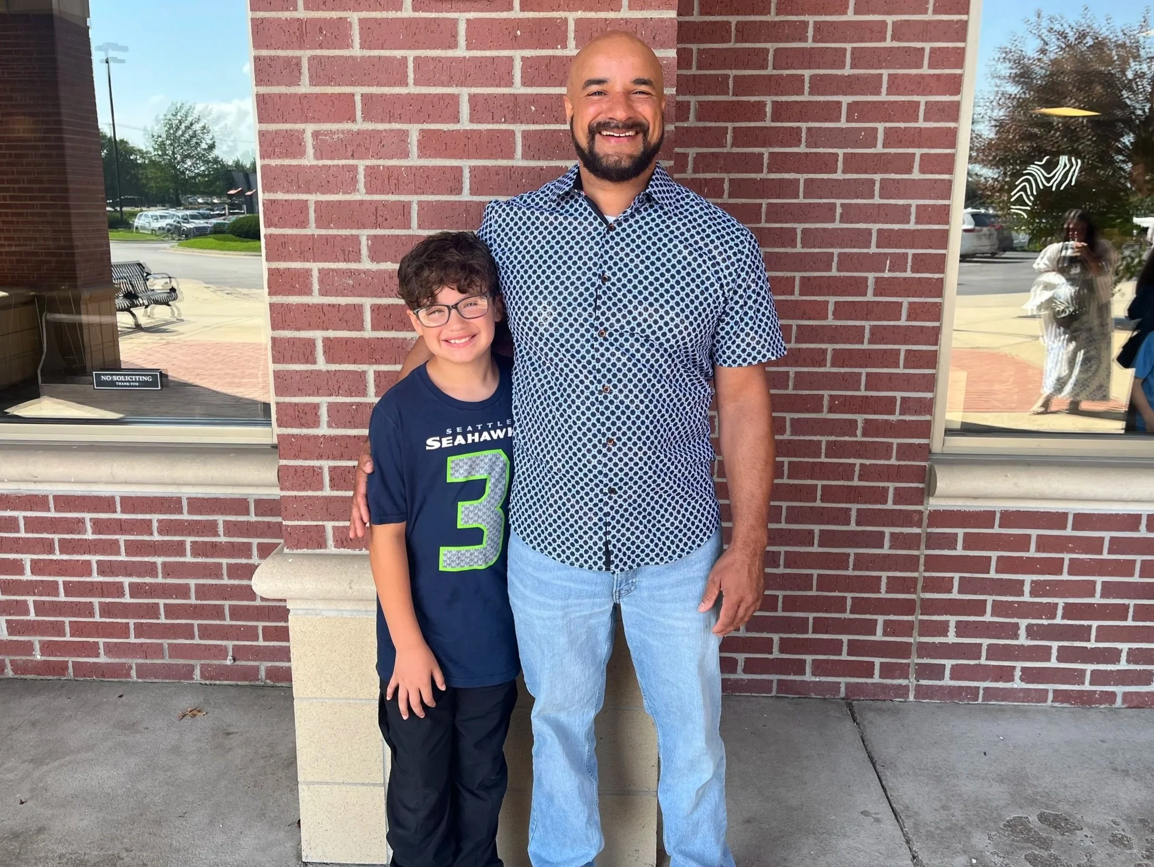A smiling man and a young boy stand together outside a red brick building