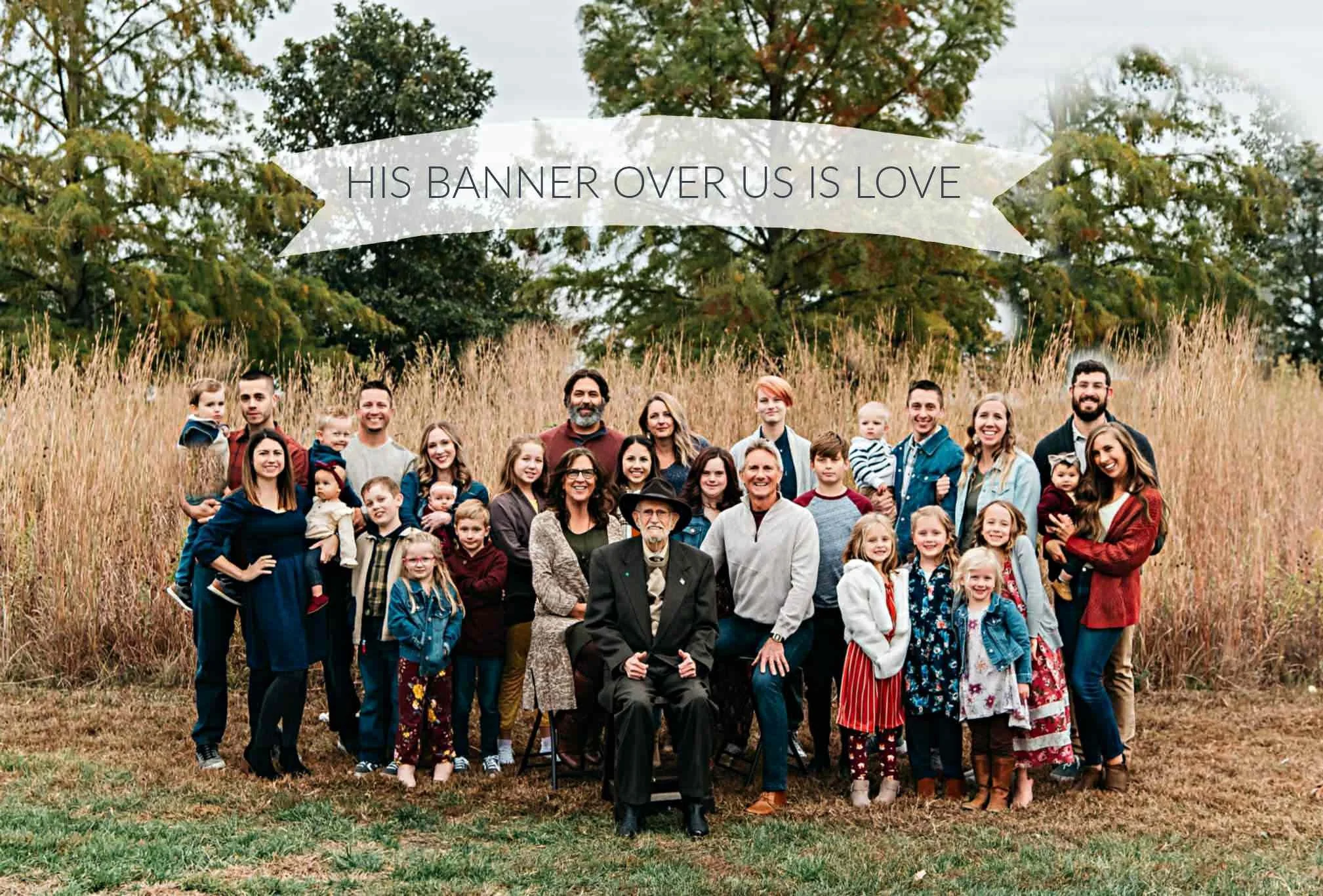 A large group of people, including children, adults, and elderly, gathered outdoors in a grassy field with trees in autumn foliage. A banner above reads 'His banner over us is love.'