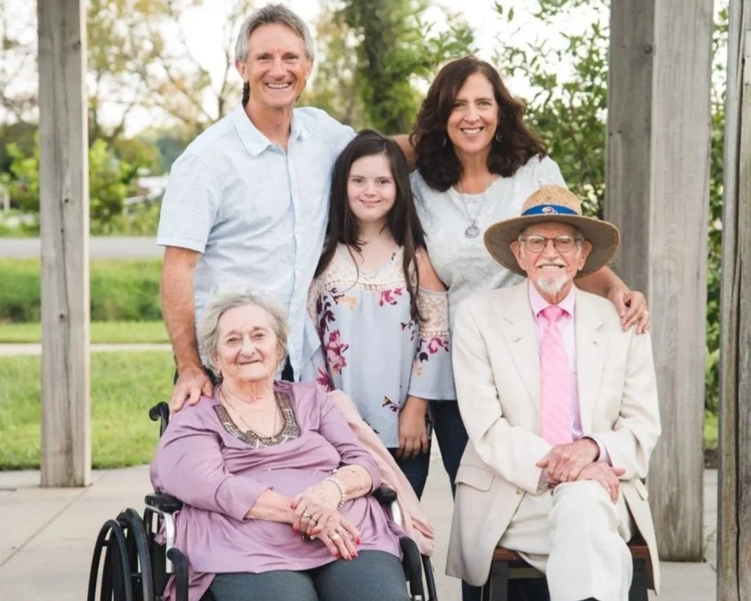 A multigenerational family portrait with three adults, a young girl, and an elderly woman in a wheelchair, outdoors with trees and a grassy area in the background.