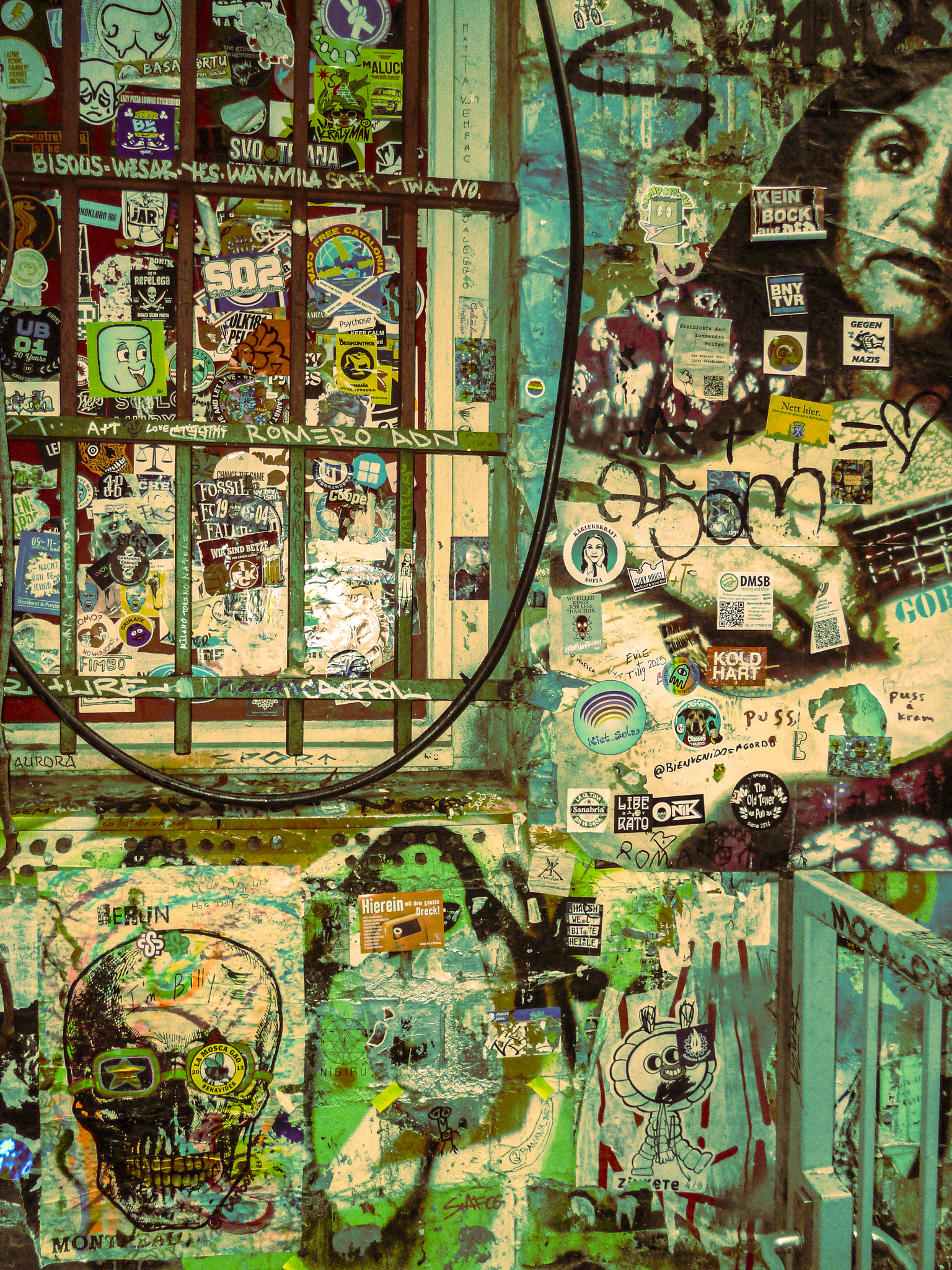 A wall covered with numerous stickers, posters, and graffiti, including a large partial portrait of a woman, various cartoon characters, skull images, and handwritten tags.