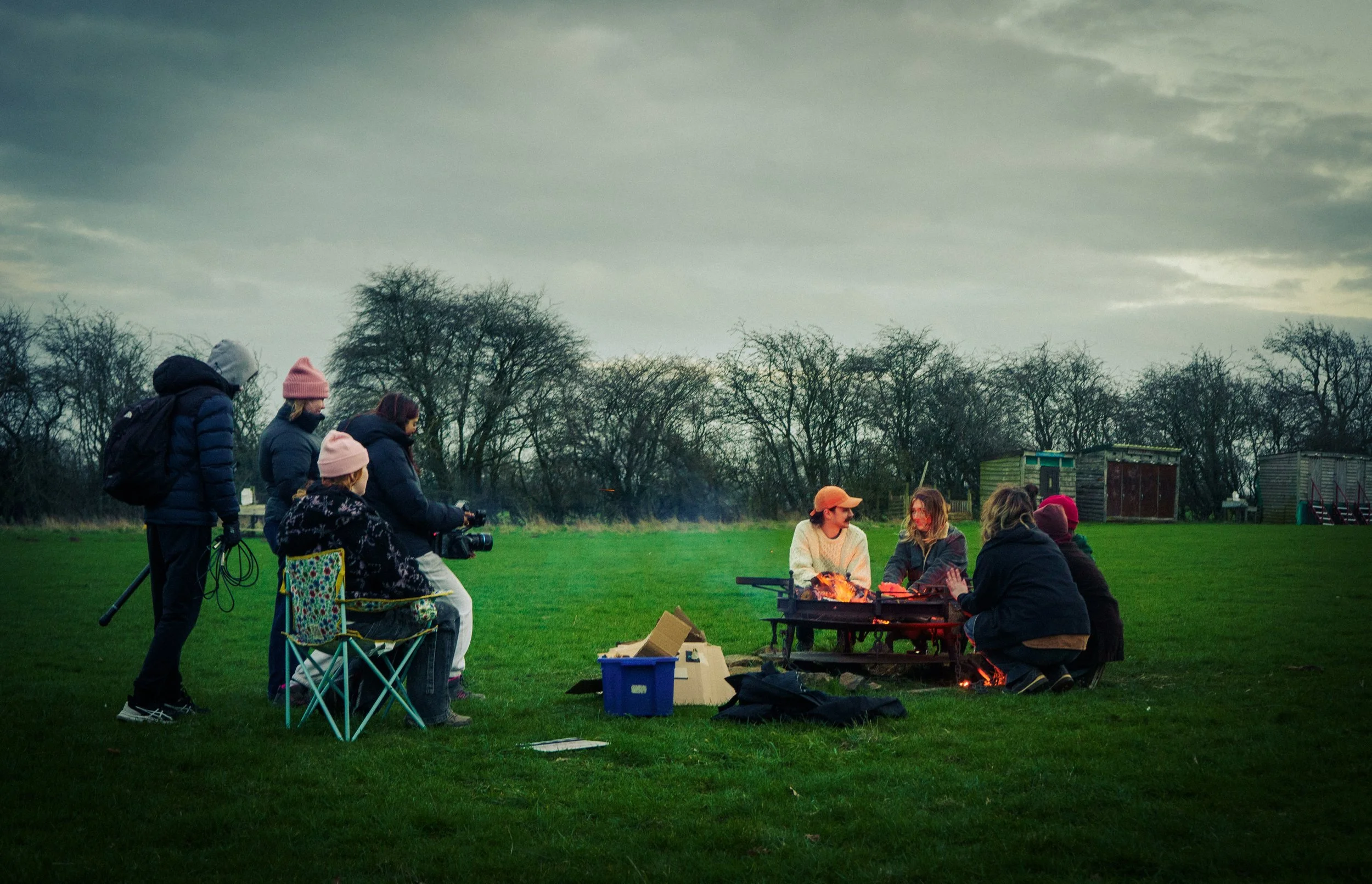 Group of people gathering outdoors around a firepit on a grassy field, some sitting on folding chairs and others standing nearby, with trees and small sheds in the background, under a cloudy sky.