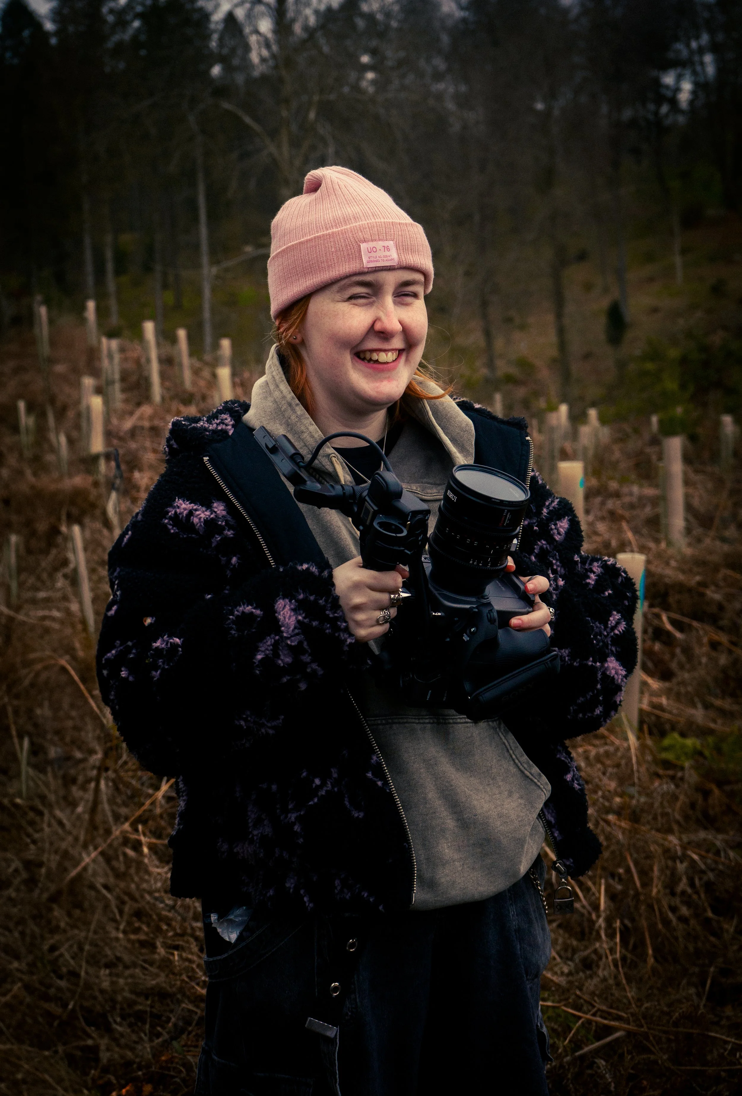A young person outdoors, smiling while holding a camera with a large lens, dressed warmly with a pink beanie, hoodie, and jacket, in a natural setting during dusk or early evening.