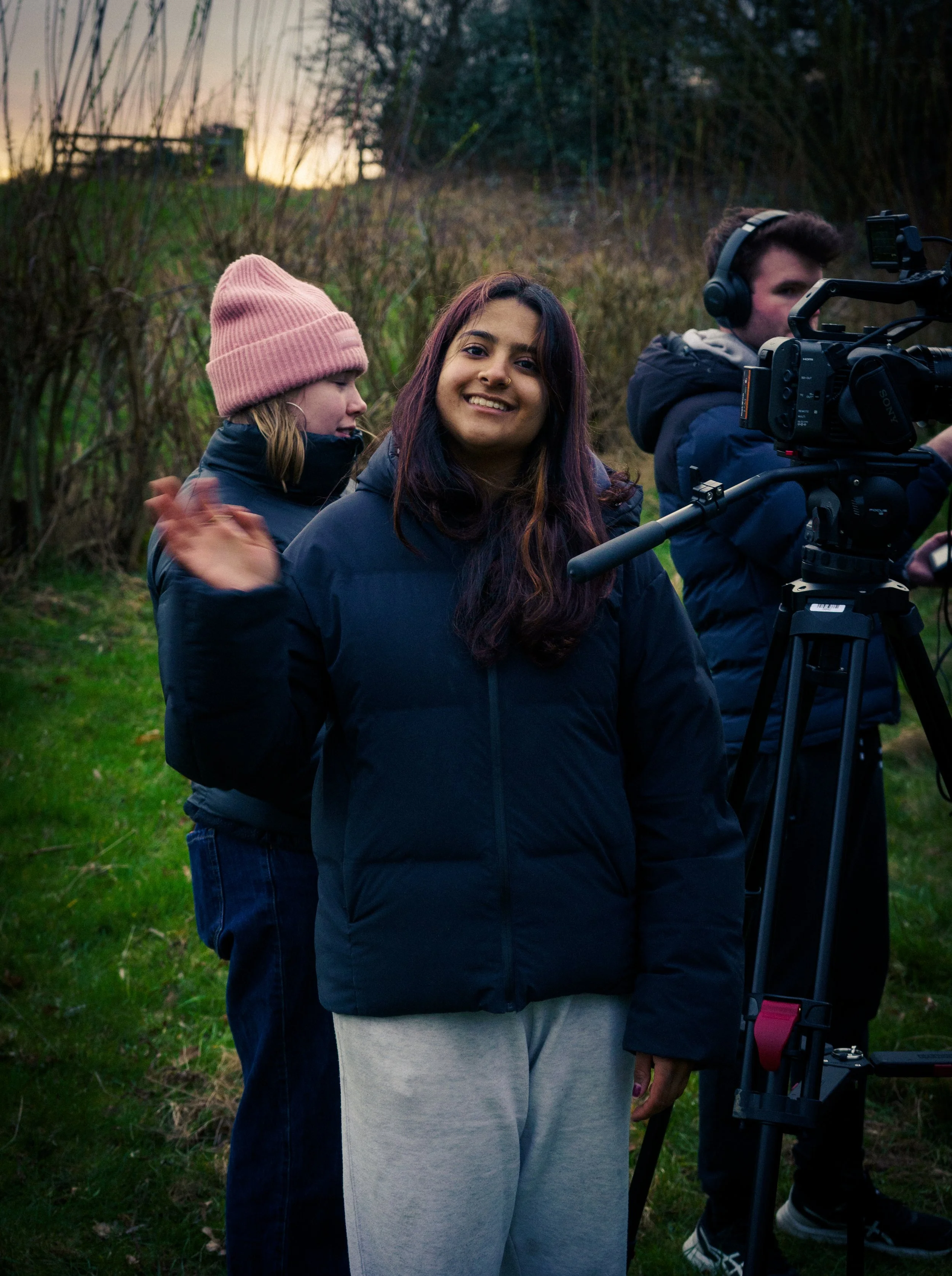 Three young people outdoors near a camera, one girl smiling and waving, others adjusting equipment, surrounded by tall grasses at sunset.