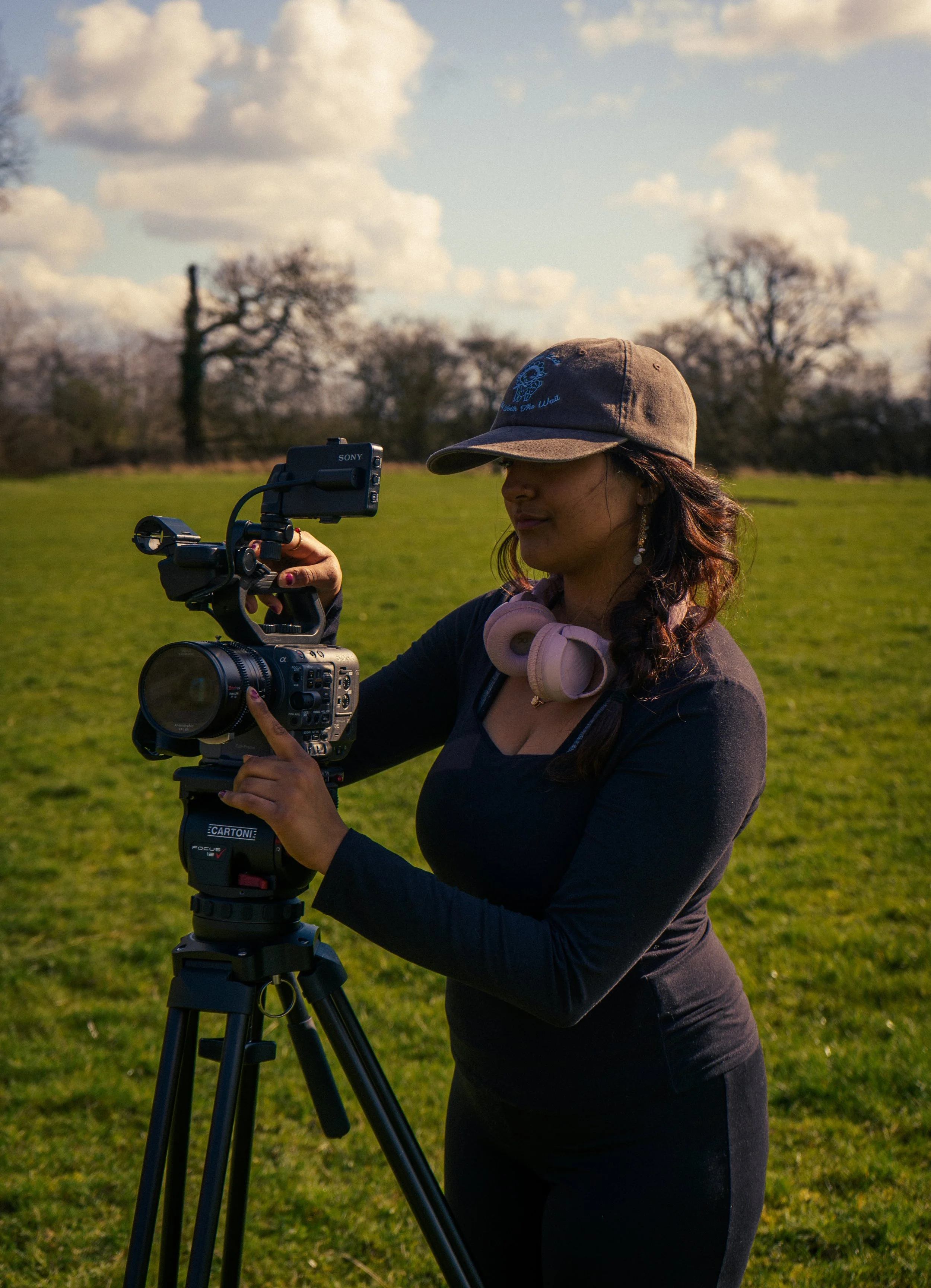 A woman setting up a video camera on a tripod in an open grassy field with trees and a cloudy sky in the background.