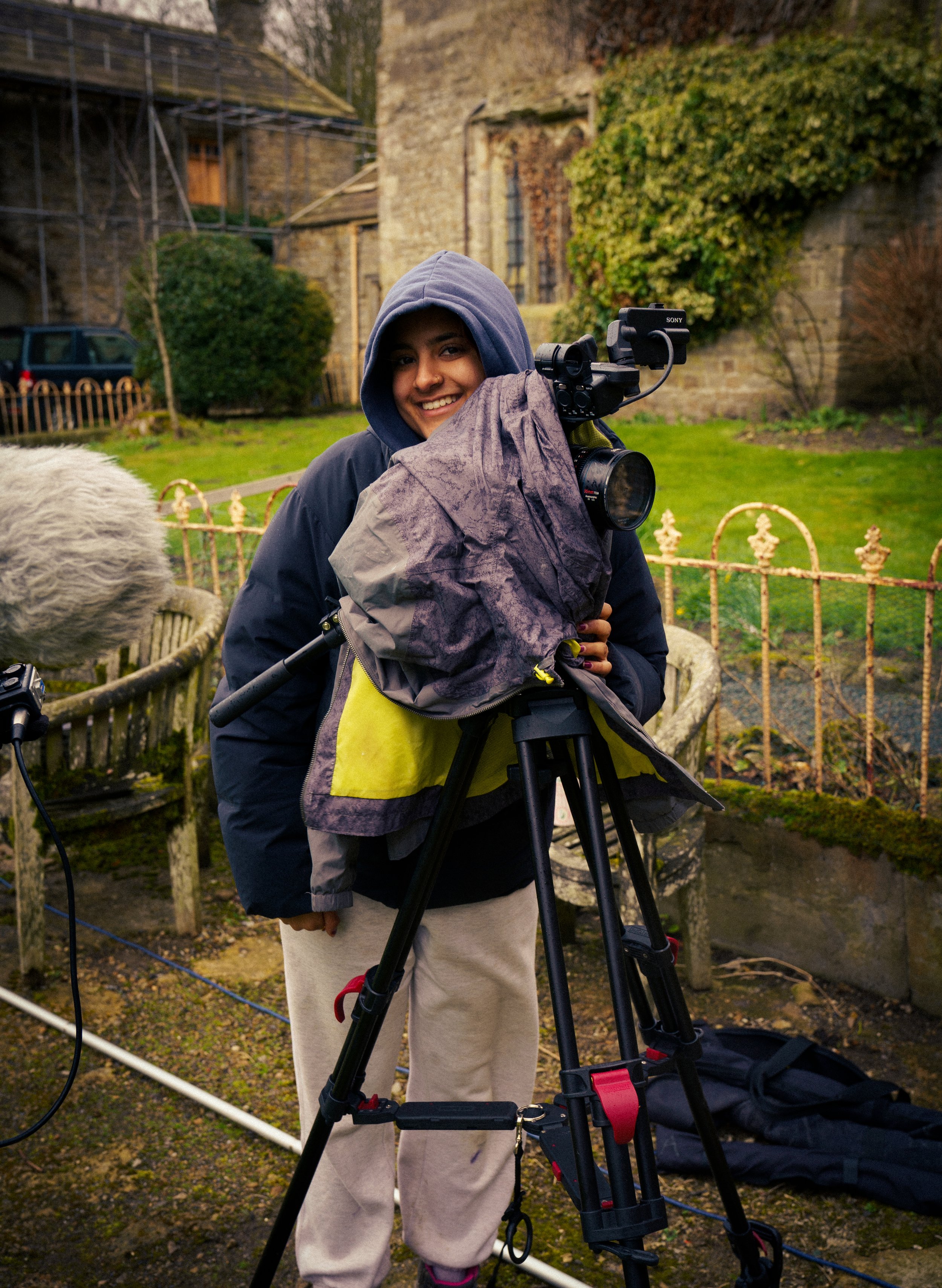 A woman in a hoodie and beige sweatpants operating a professional camera on a tripod outdoors, smiling at the camera with greenery and old stone buildings in the background.