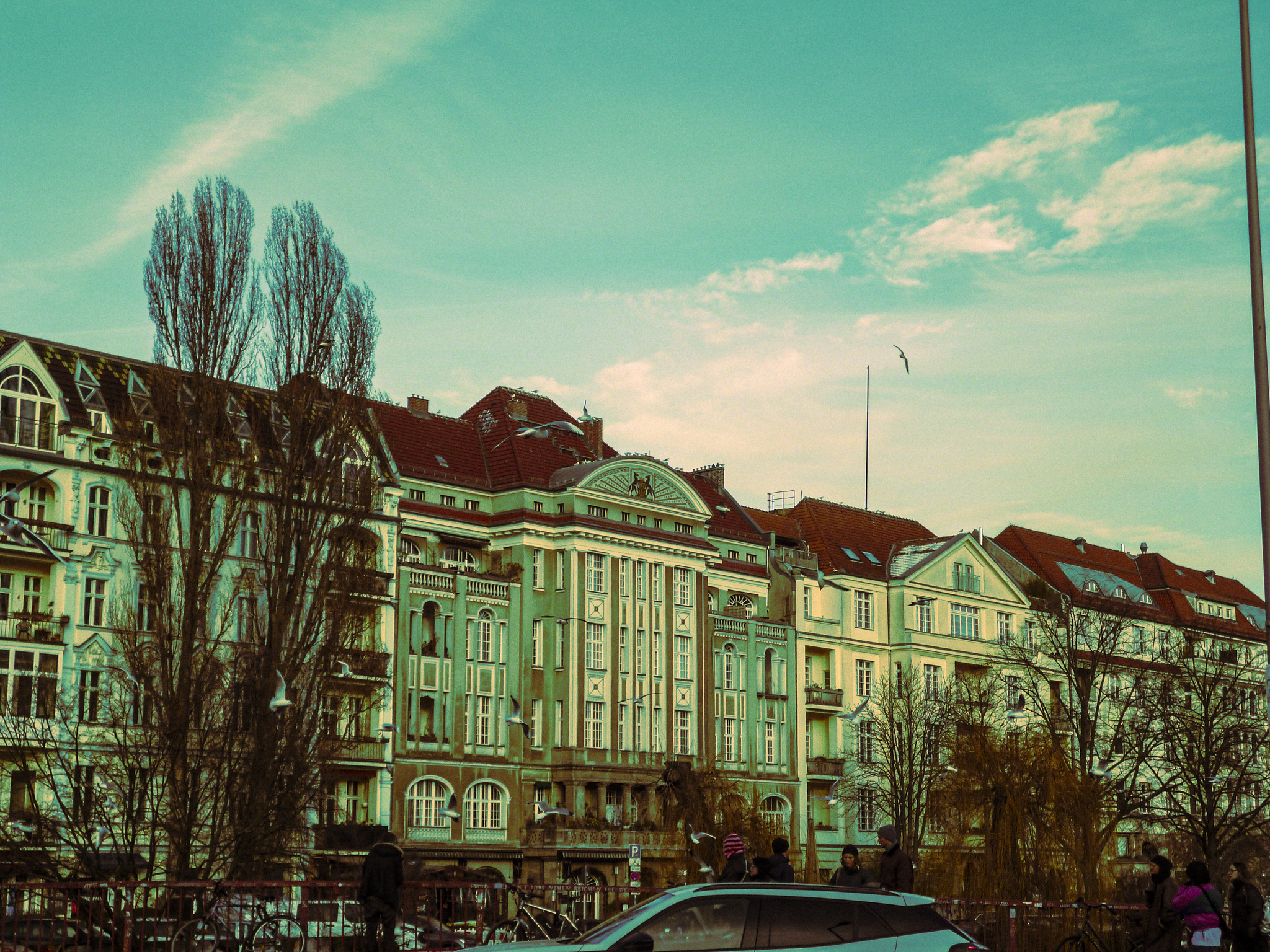 City street view with historic buildings, leafless trees, birds flying, and people walking near parked cars under a sky with some clouds.