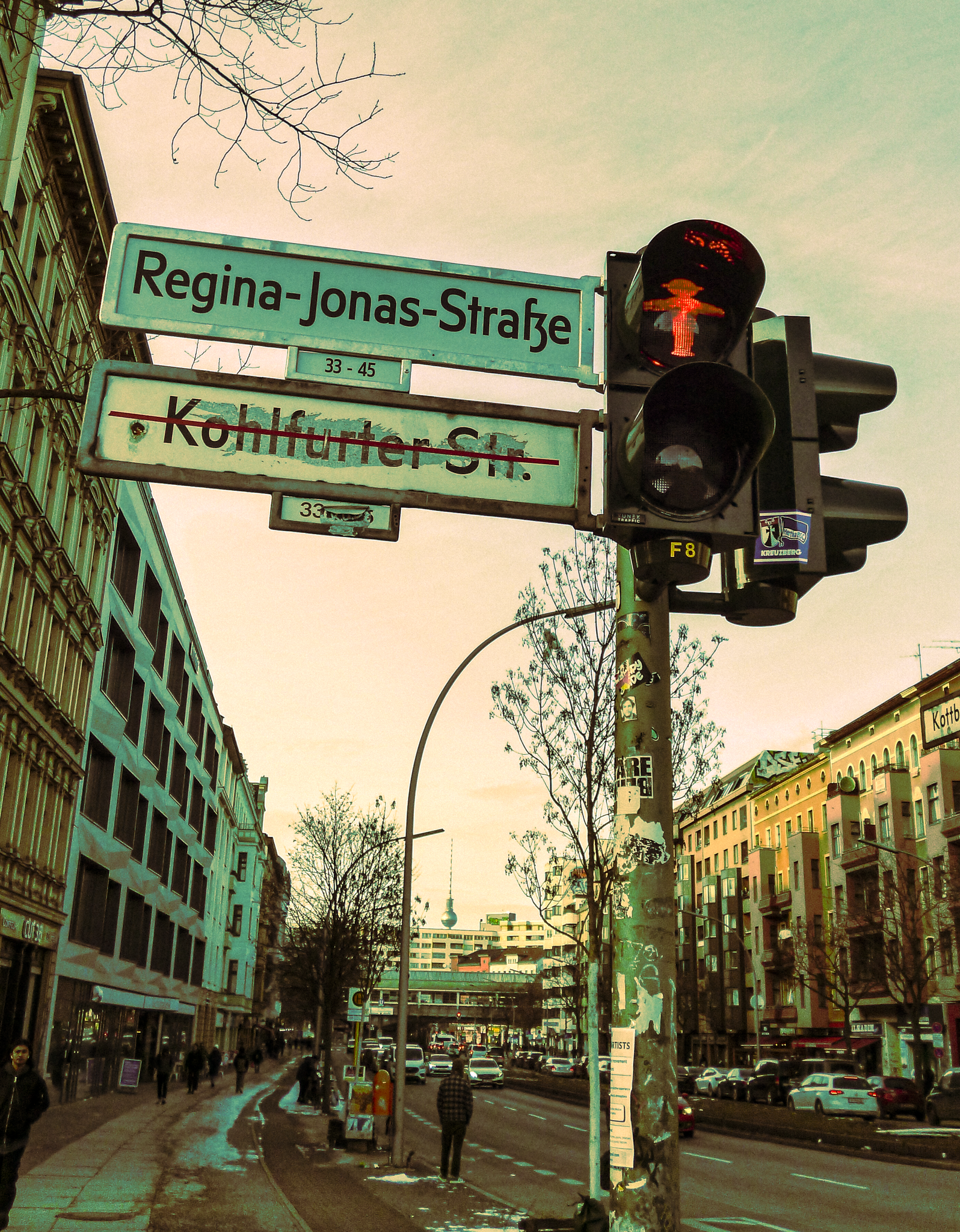 Street sign showing 'Regina-Jonas-Straße' with a traffic light and surrounding buildings in an urban area, with a TV tower in the background.