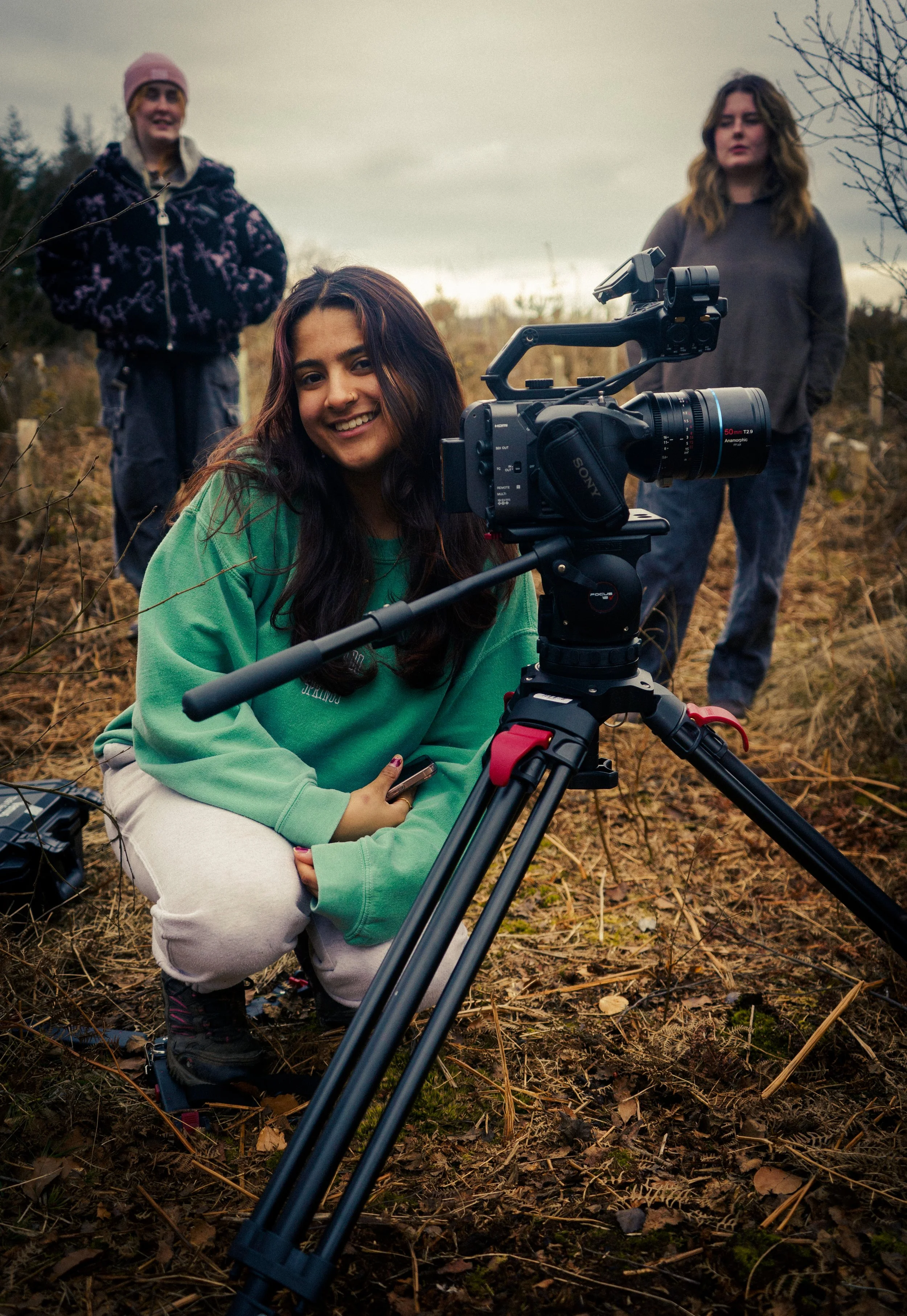 Three young people in outdoor setting with dry grass and trees, one smiling while kneeling and operating a video camera on a tripod, the others standing in the background under a cloudy sky.