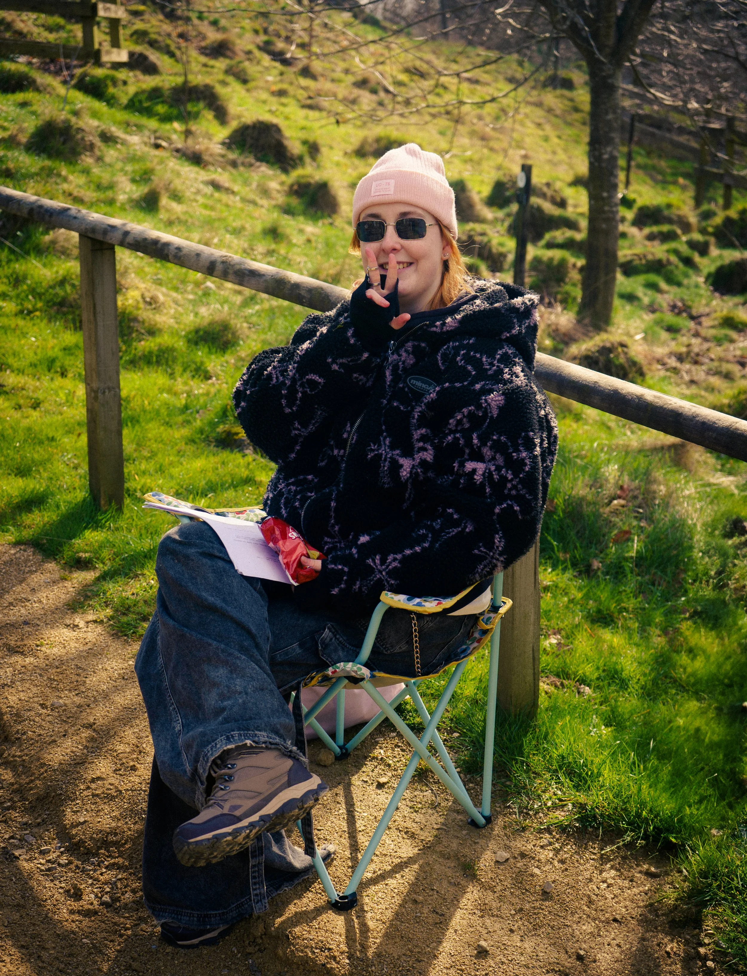 A person sitting on a folding camp chair outdoors in a grassy area, wearing a pink beanie, sunglasses, a black and pink patterned jacket, and jeans, smiling and making a peace sign with their fingers.