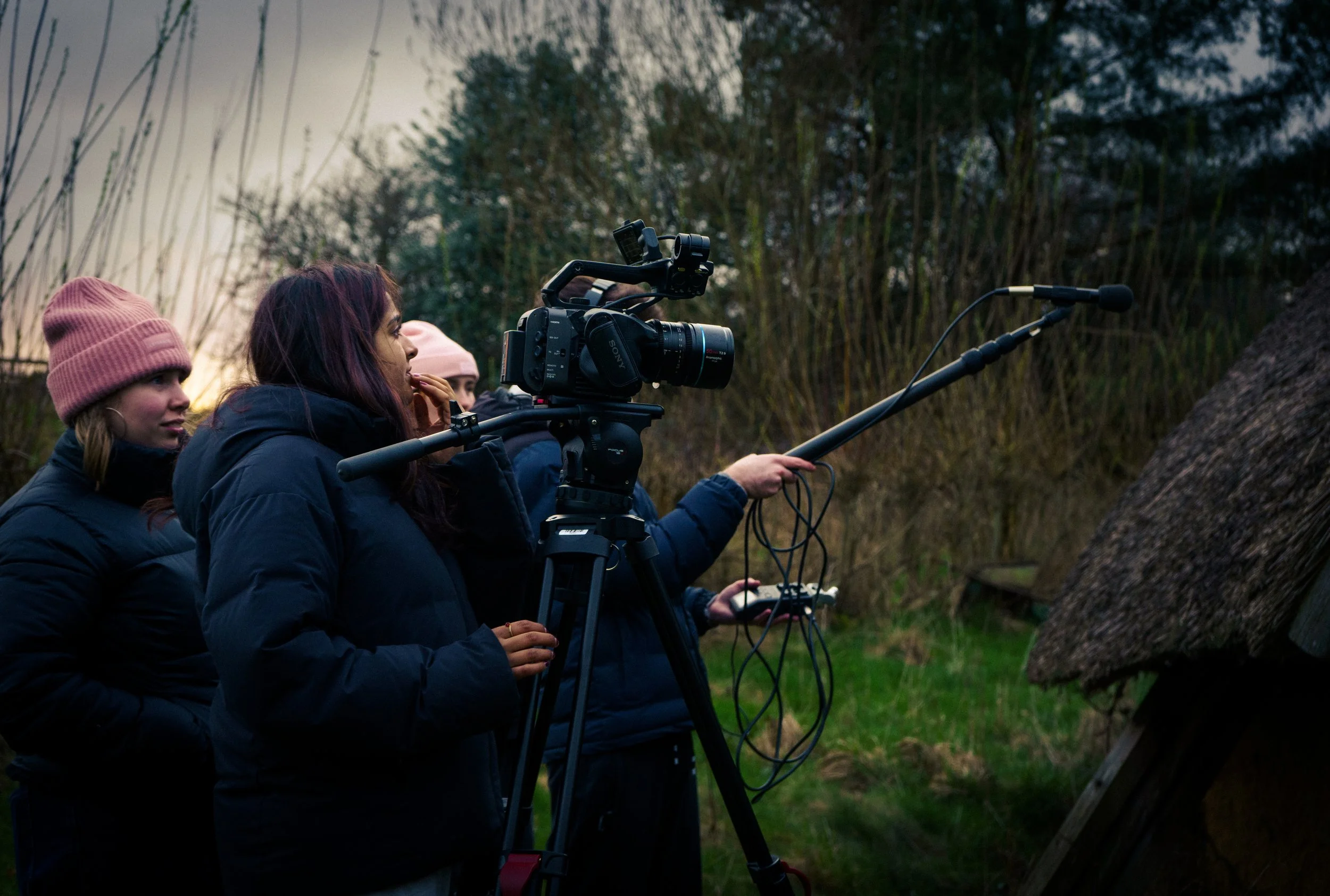 Three people involved in filming outdoors, one woman operates a camera on a tripod, another woman holds a long boom microphone, and a third person observes, with trees and a thatched roof structure in the background.