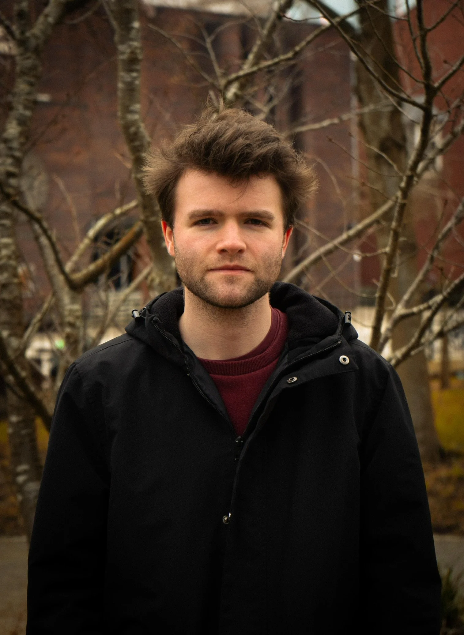 Young man with dark brown hair and beard standing outdoors in front of leafless trees and a brick building.