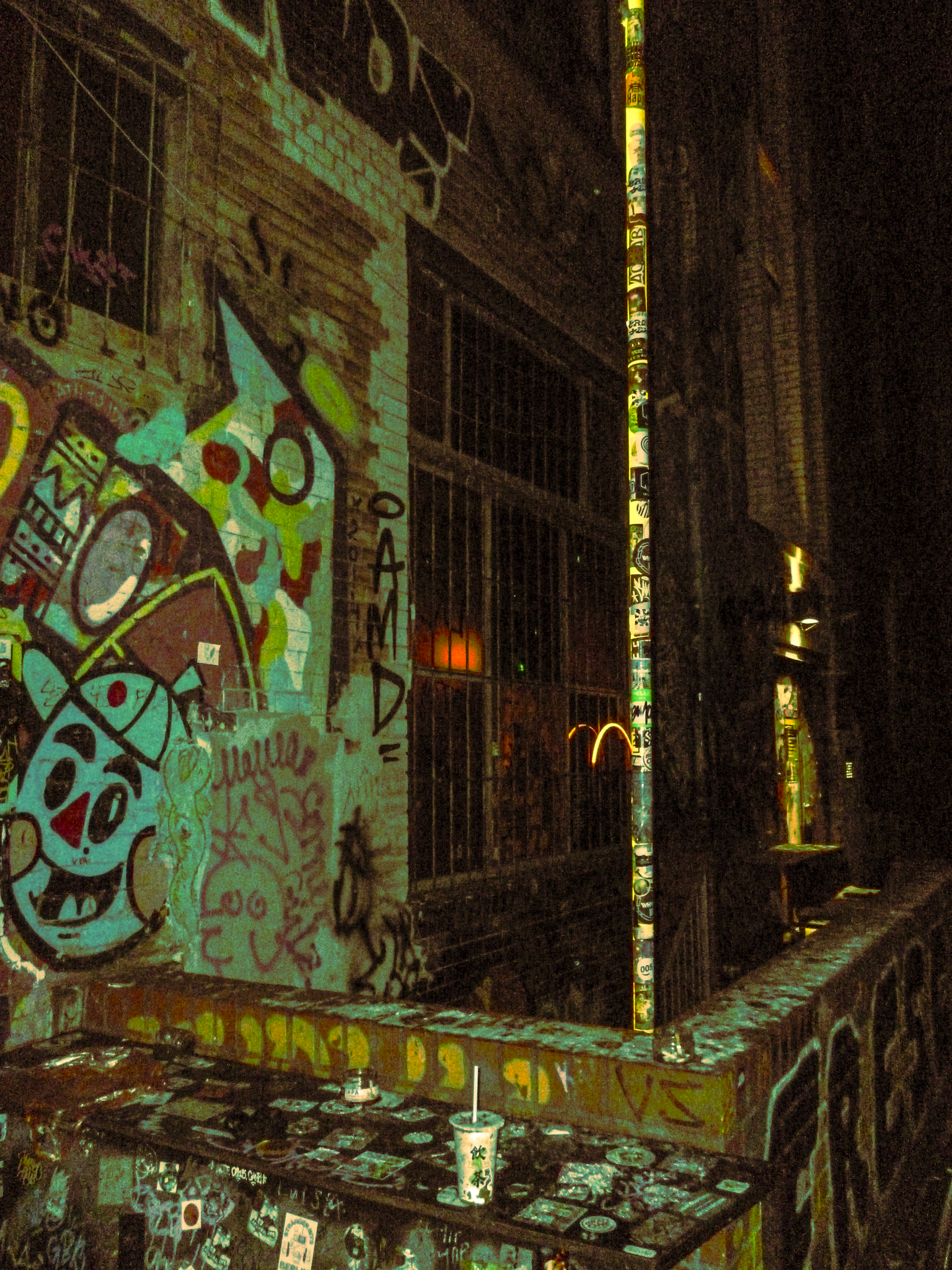 Nighttime urban scene featuring graffiti and street art on brick walls, metal bars on windows, a vertical column covered in stickers, and a small table with stickers and a drink cup.