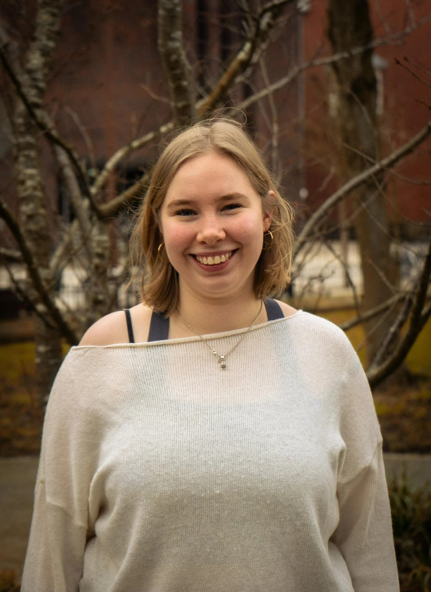 A woman smiling outdoors in front of a tree with bare branches, wearing a beige sweater, a black tank top, and jewelry.