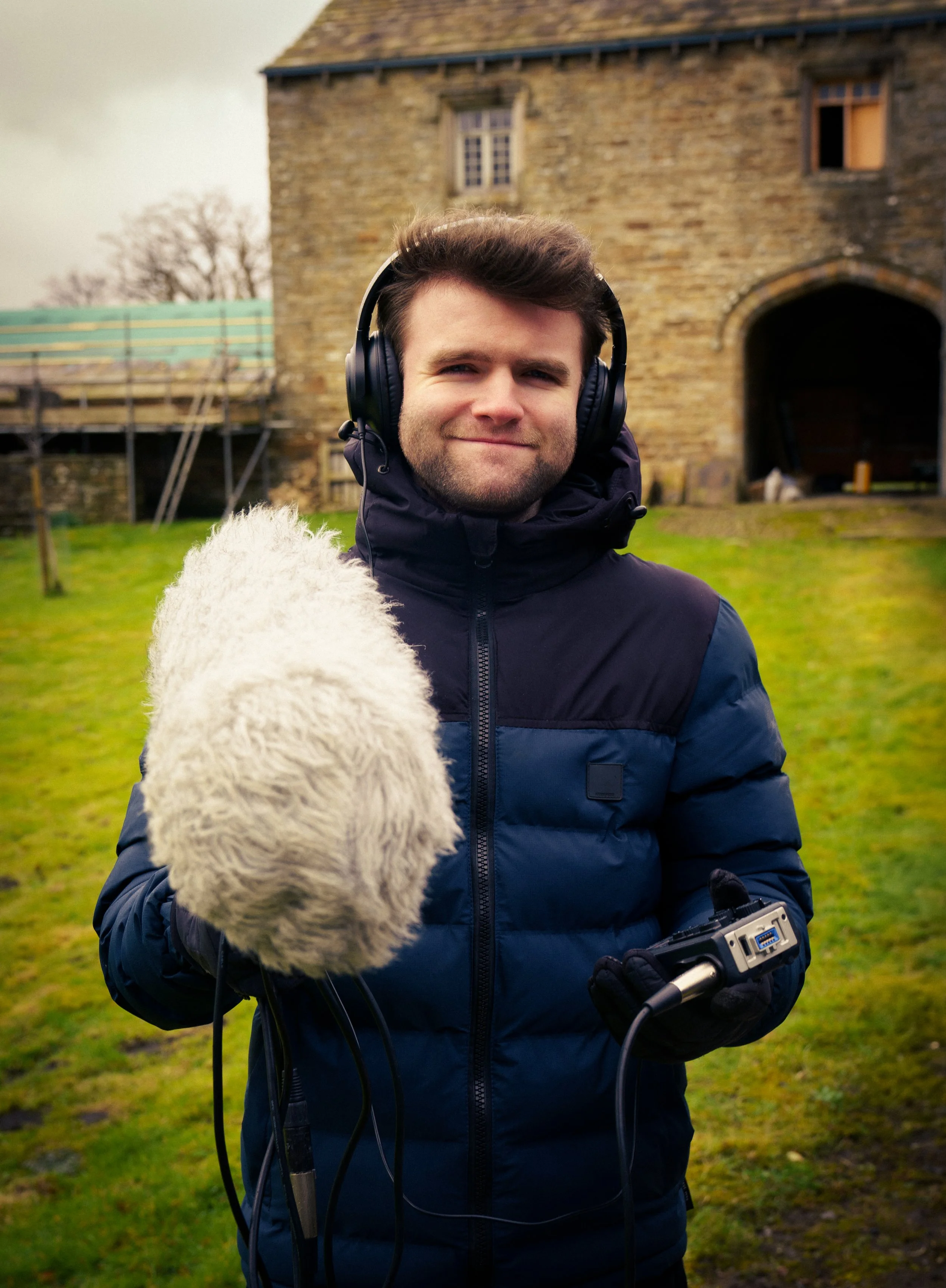 A man outdoors wearing headphones, holding audio recording equipment and a microphone, with a stone building and grassy area in the background.