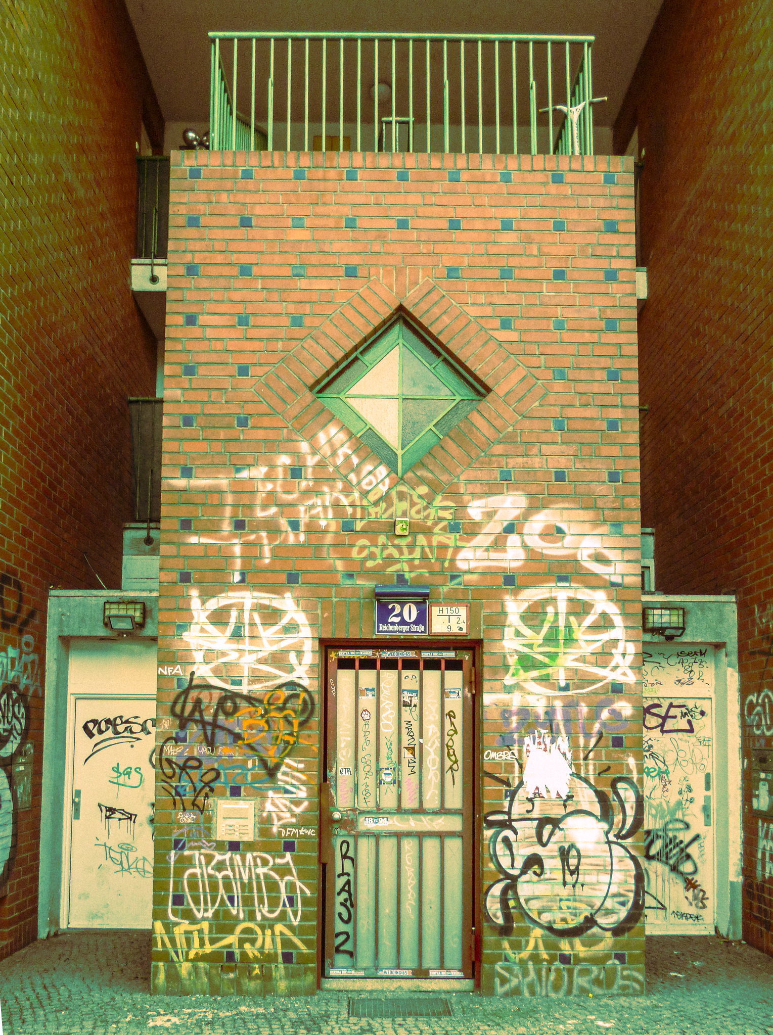A brick building with graffiti, featuring a central metal door, flanked by two white doors also covered in graffiti. The building has a diamond-shaped green window in the upper center and a green railing on the upper balcony.