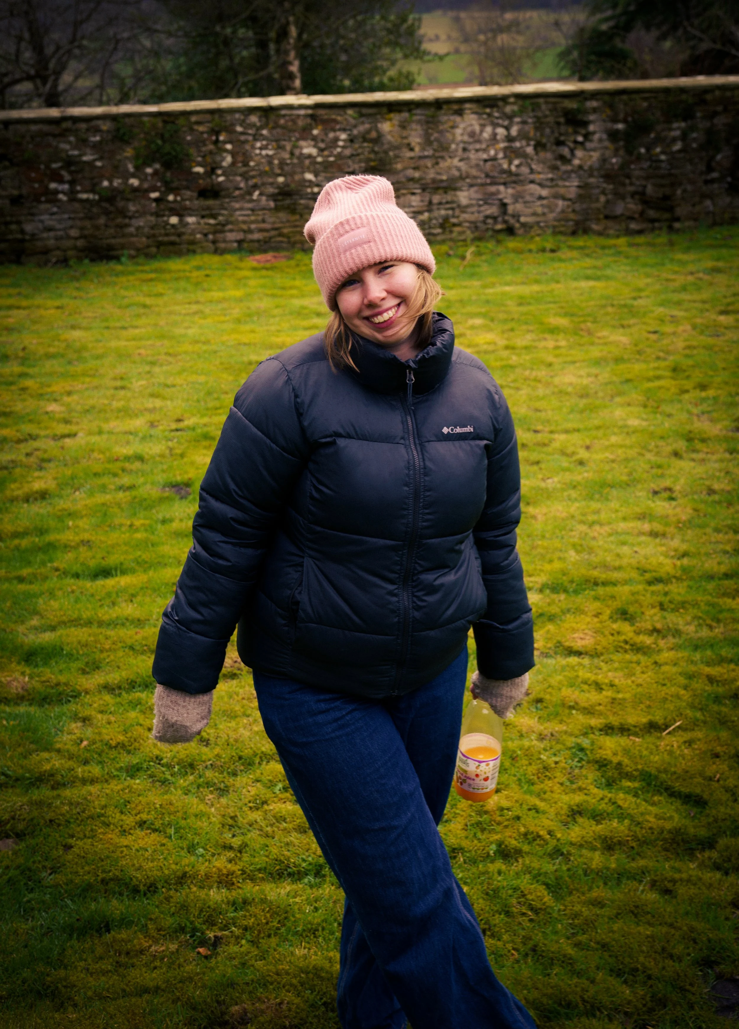 A young woman smiling outdoors on a grassy field, wearing a pink knit hat, a black puffy jacket, and jeans, holding a clear bottle of orange juice.