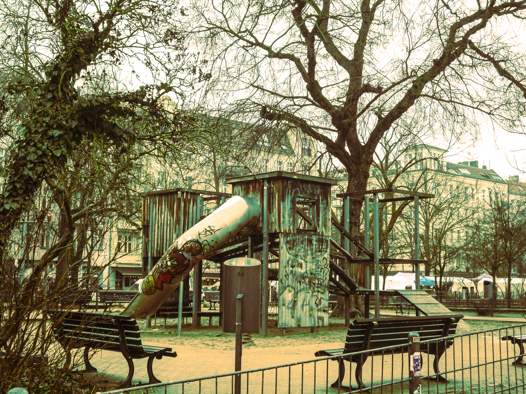 A weathered wooden playground structure with a slide and climbing walls in a park, surrounded by leafless trees and benches.