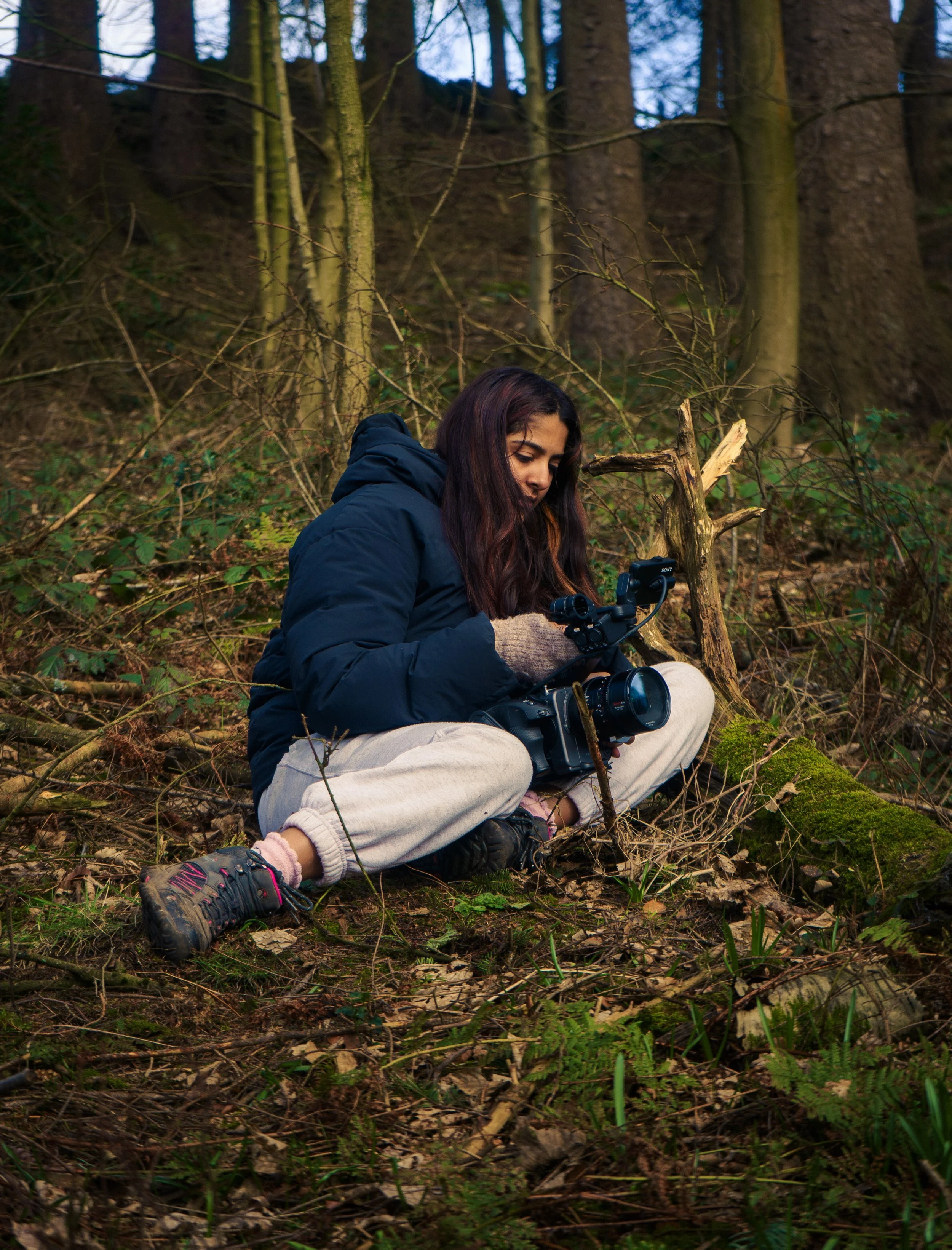 A woman with dark hair sitting cross-legged on the forest floor, holding a professional camera, surrounded by trees and foliage.