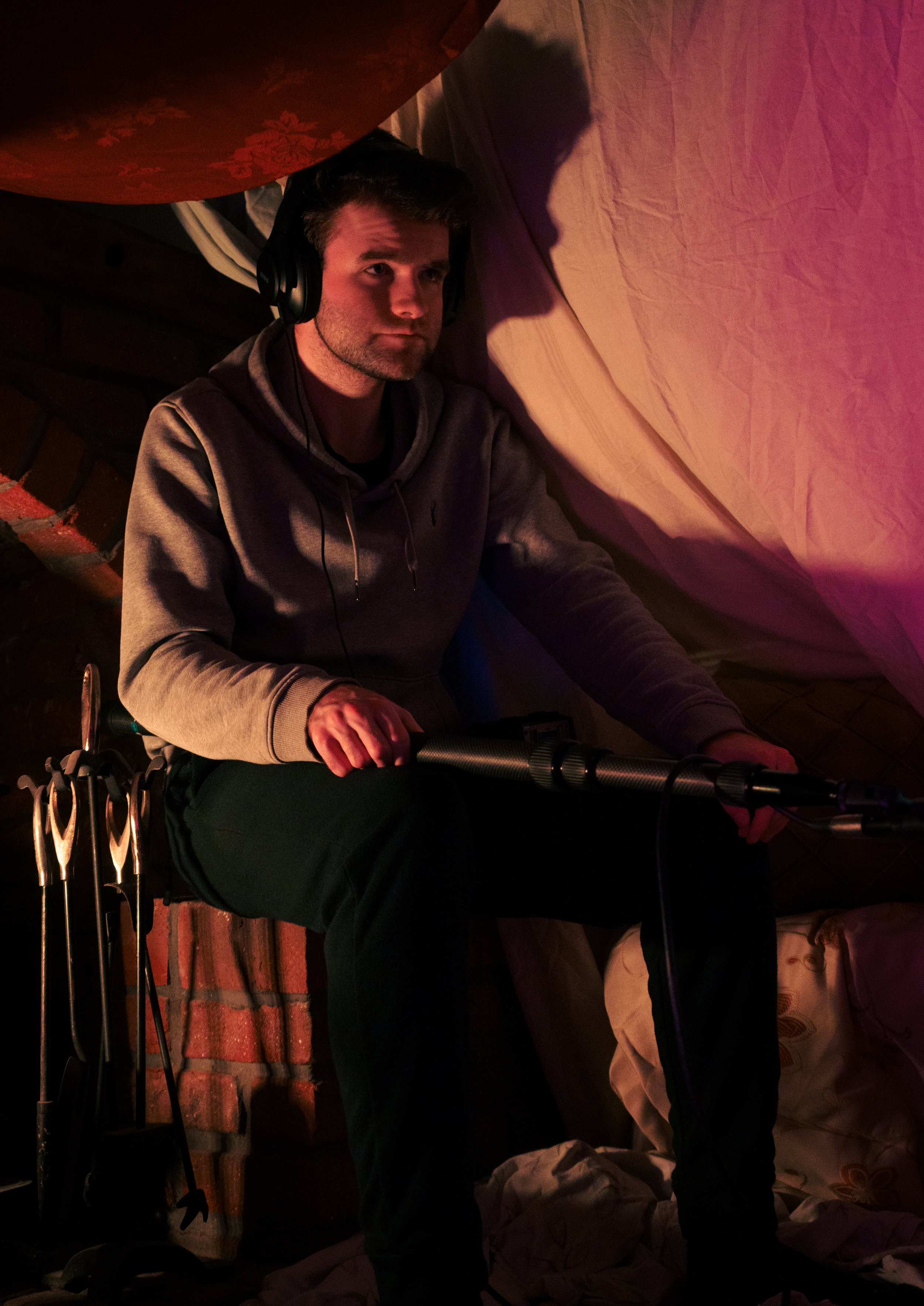 A man with headphones sitting on a chair, surrounded by blankets and pillows, in a cozy indoor space with soft lighting.