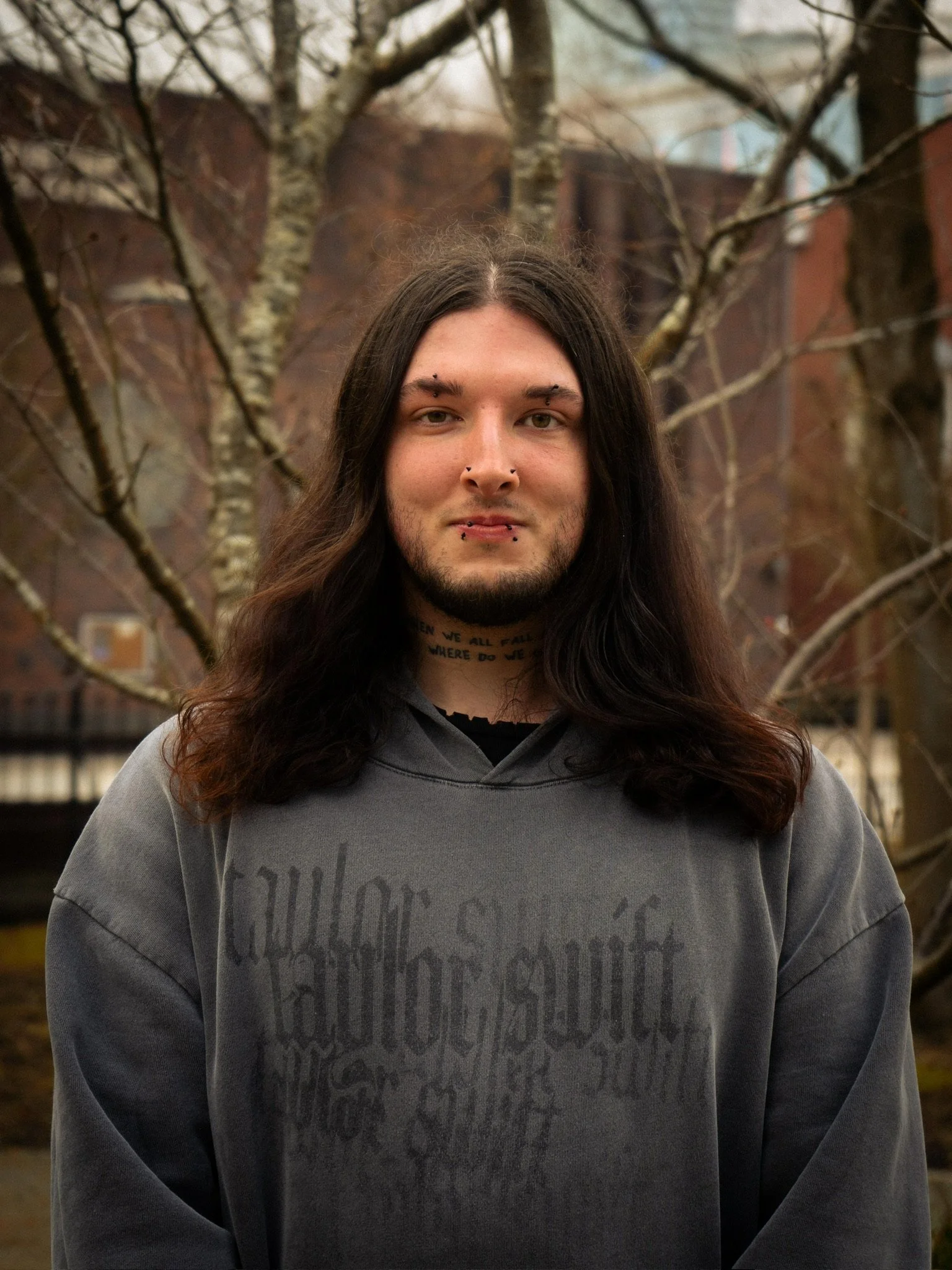 A young man with long dark hair, facial piercings, and tattoos, standing outdoors in front of a leafless tree and brick buildings.